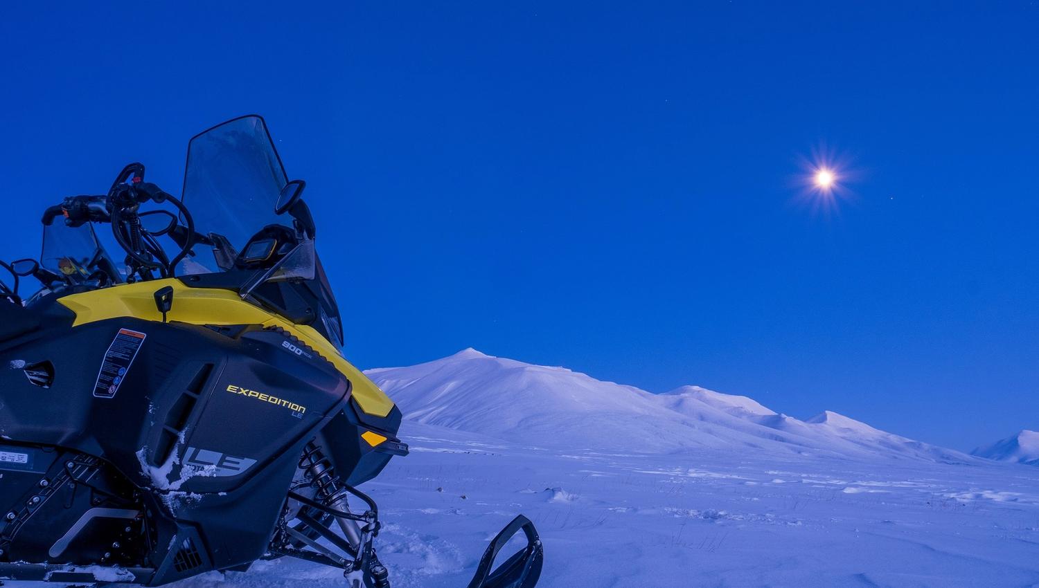 A snowmobile in a snow-covered landscape covered in blue light with a bright moon in the sky