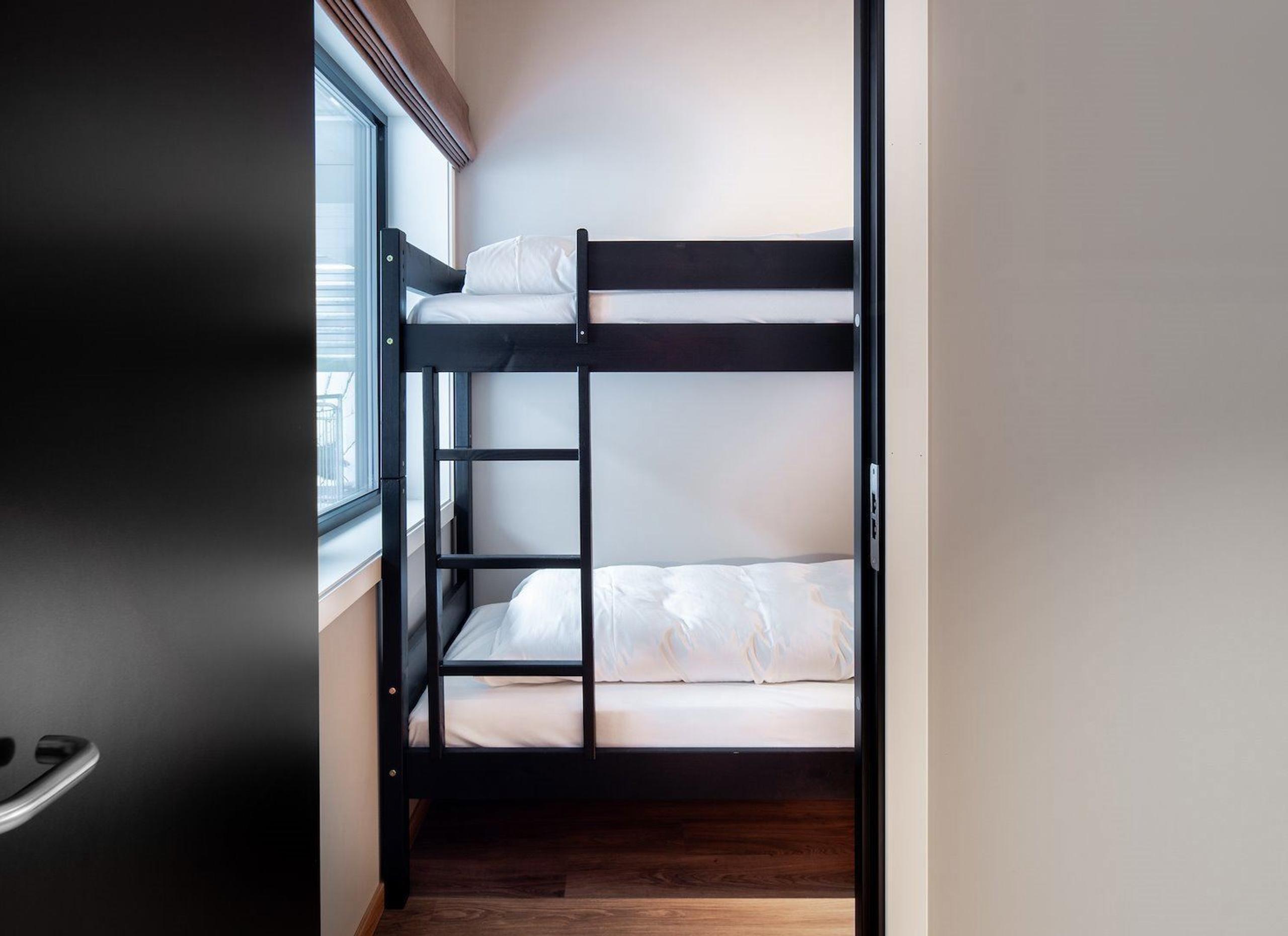 Black wooden bunk bed with white bedding in a narrow room, beside a large window and wood floor.