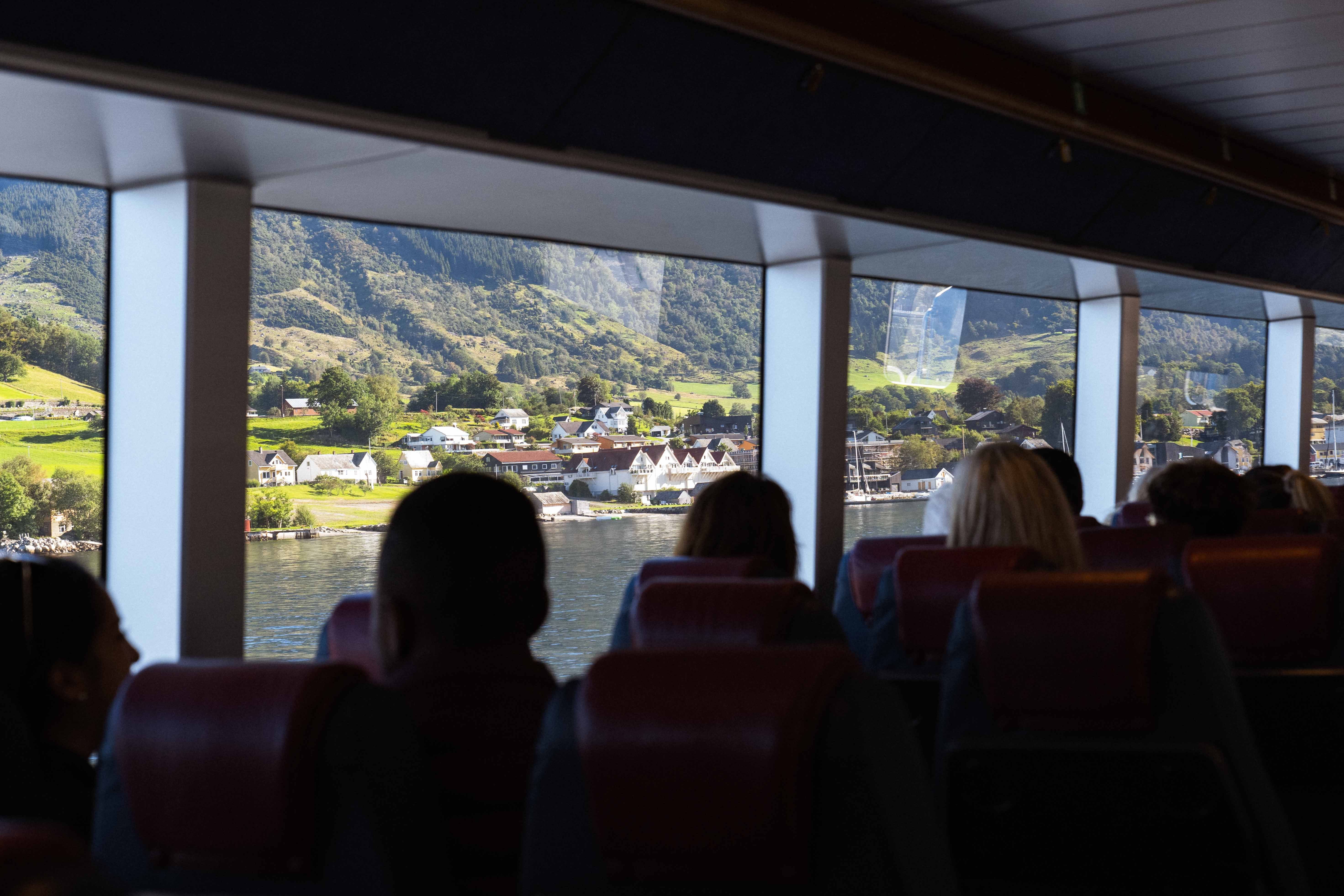 Passengers enjoying the fjord view from inside Hardangerfjordekspressen, with a village and mountain landscape in the background.