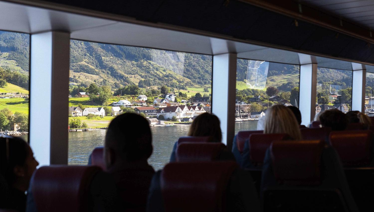 Passengers enjoying the fjord view from inside Hardangerfjordekspressen, with a village and mountain landscape in the background.