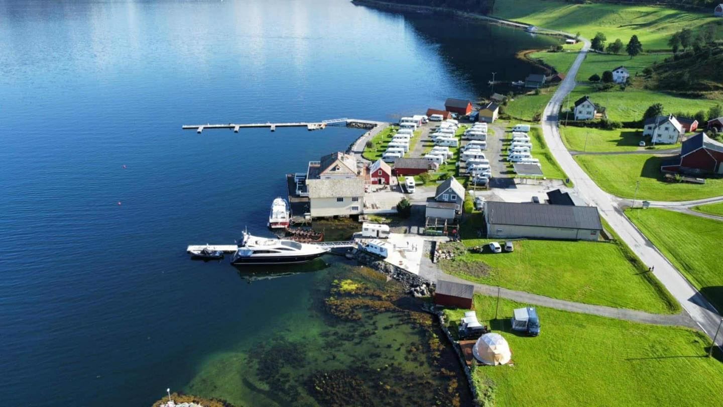 Aerial view of Davik Fjordparadis in Bremanger - a lakeside marina with docks, boats, and a campground of RVs beside green fields and a road..