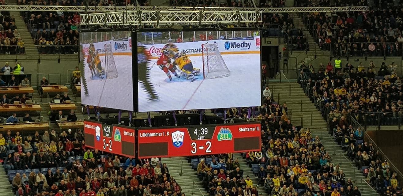 Publikum fyller tribunene i en innendørs ishockeyarena, med storskjerm som viser kamp og resultattavle..