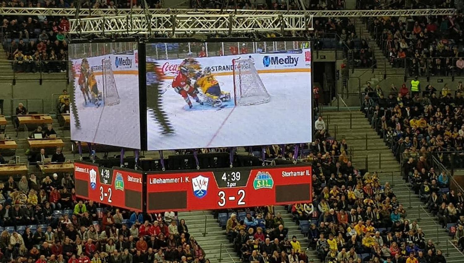 Publikum fyller tribunene i en innendørs ishockeyarena, med storskjerm som viser kamp og resultattavle..