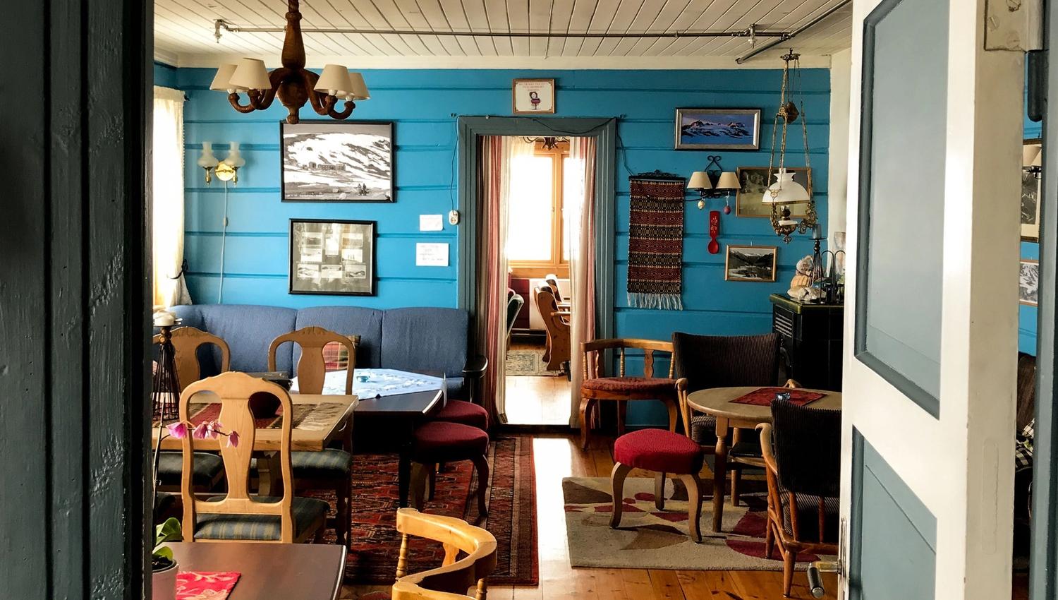 Vintage sitting room with blue wood walls, framed photos, wooden chairs and tables, and a bright doorway to another room..