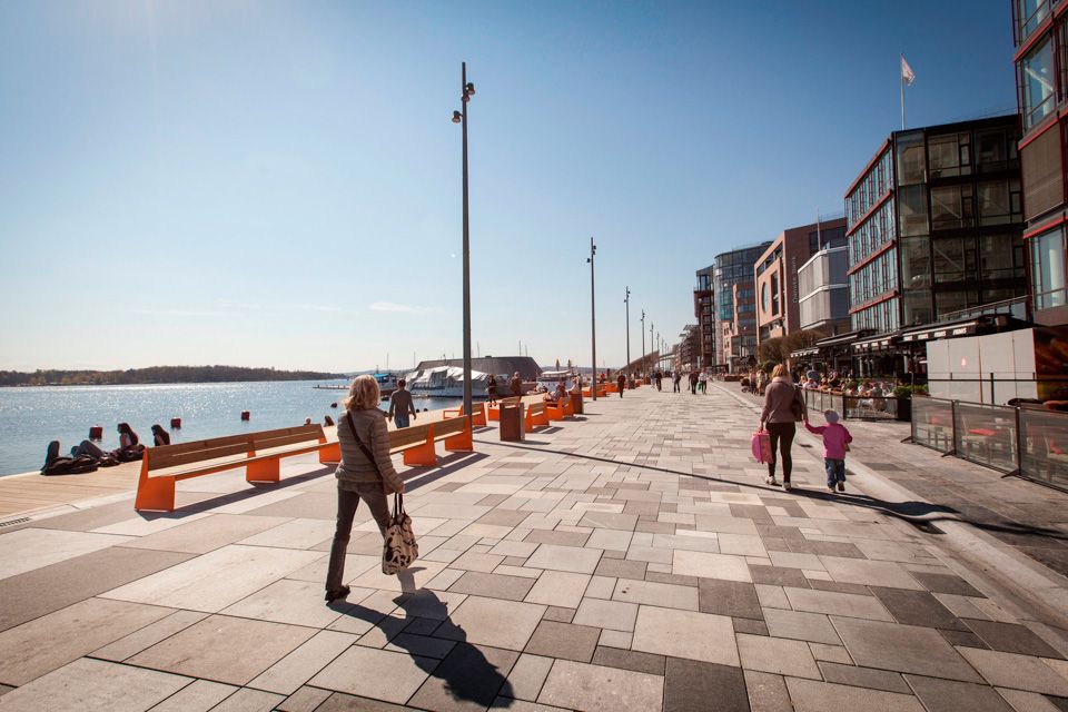 Blue sky above the promenade with people walking along it. Brown buildings with outdoor serving on the right-hand side.