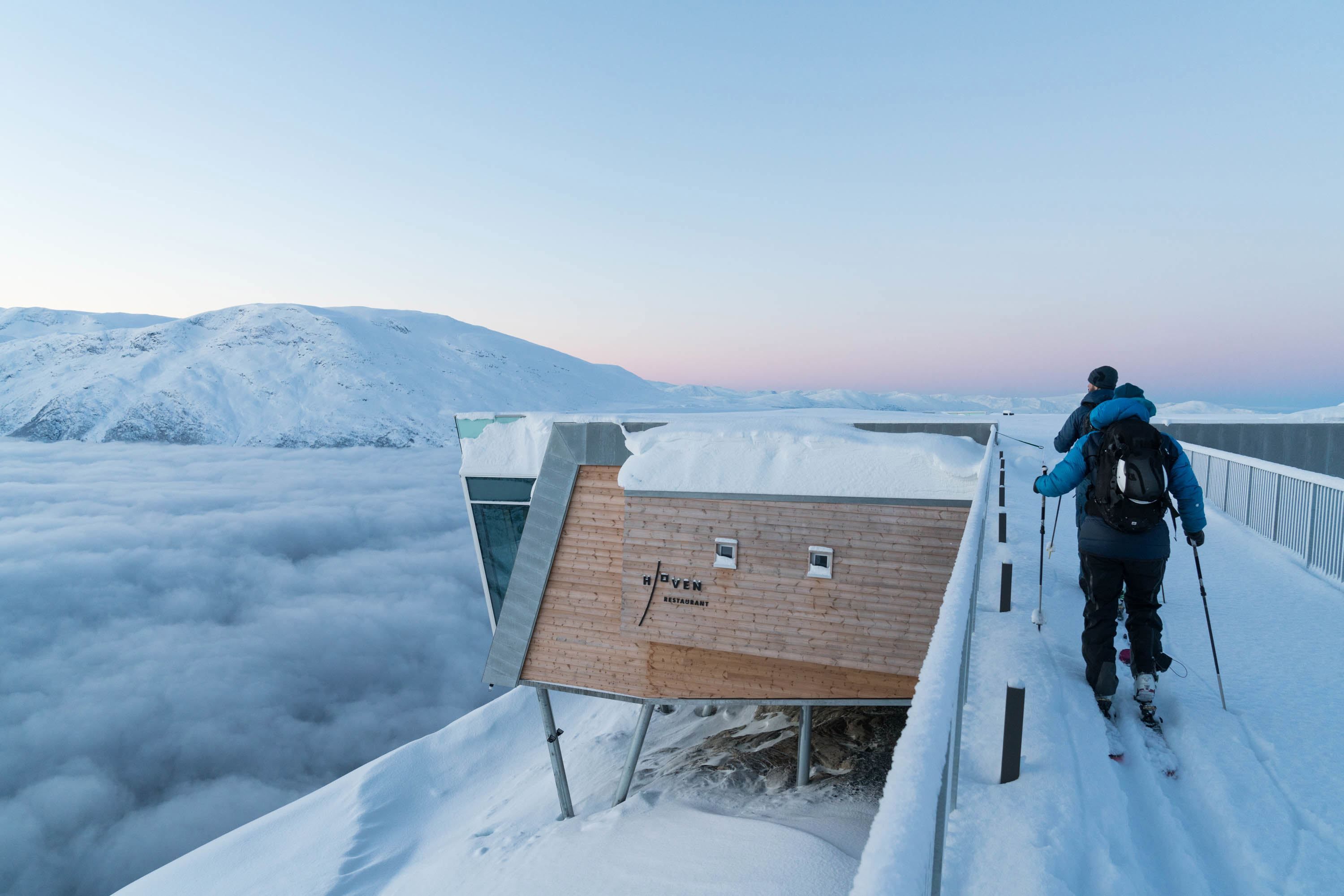 Two hikers walk along a snowy walkway beside a wood building on stilts above clouds, with mountains in the distance.