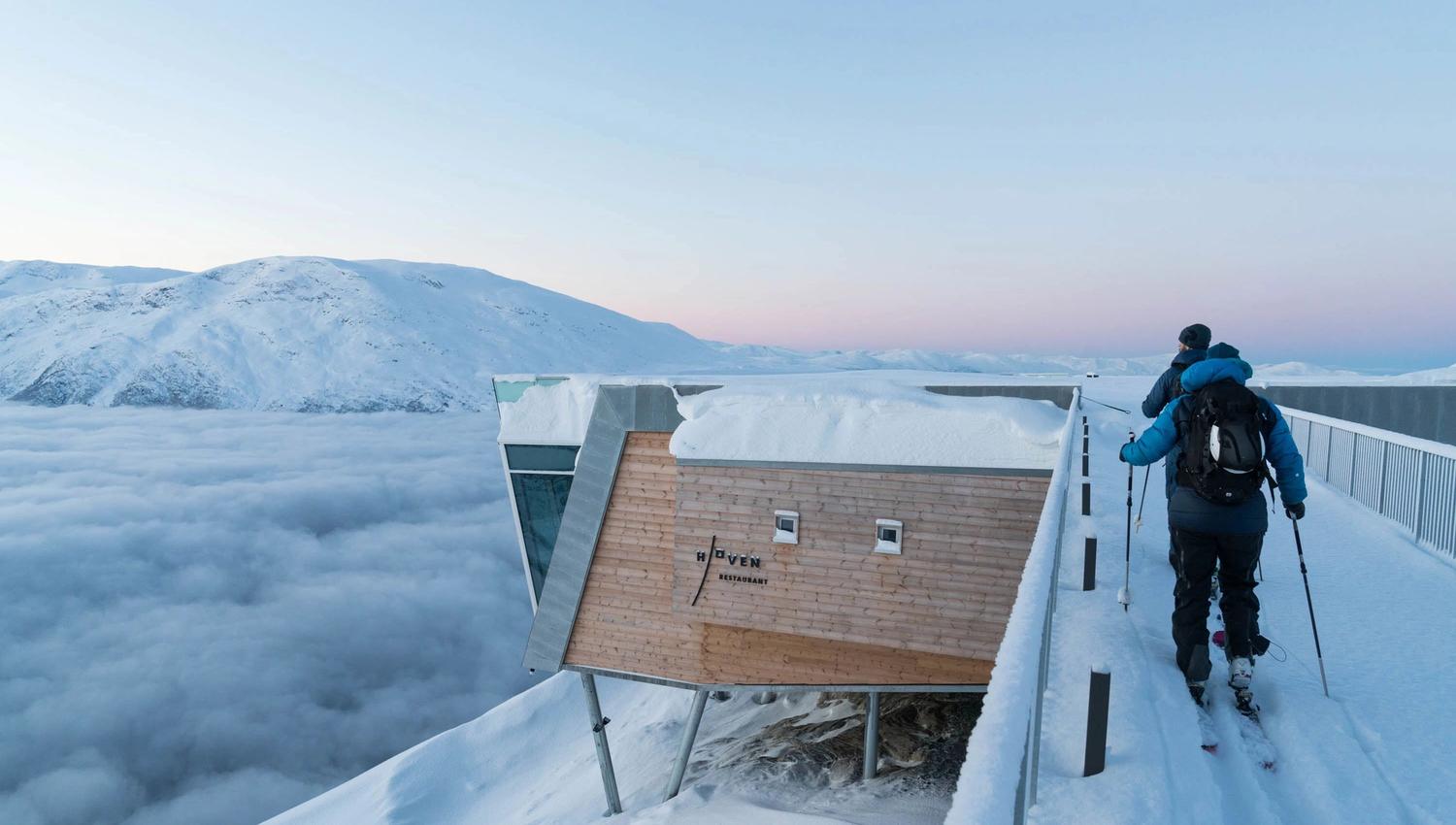Two hikers walk along a snowy walkway beside a wood building on stilts above clouds, with mountains in the distance.