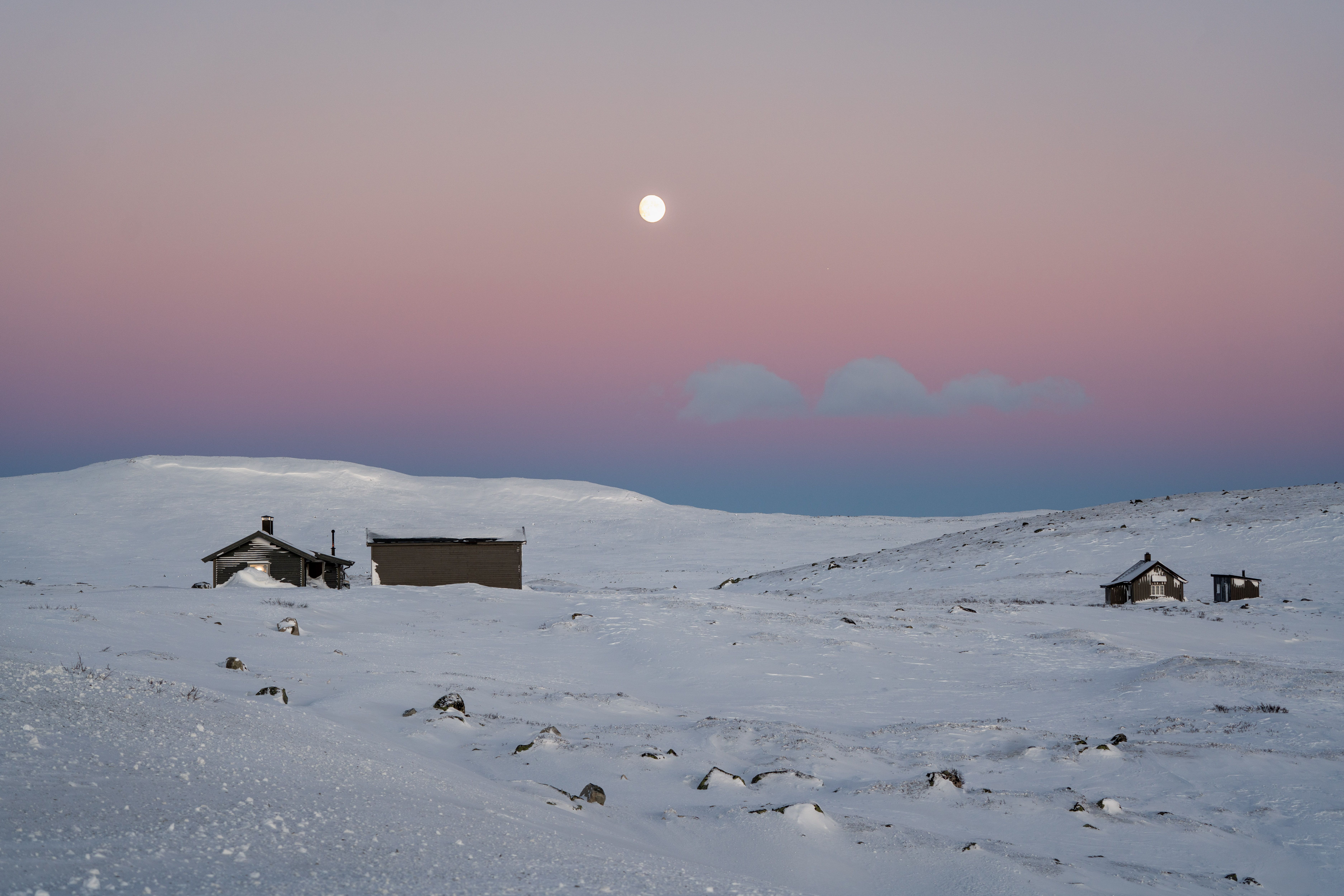 Some mountain cabins in the winter landscape underneath a pink sky with a full moon in the background.