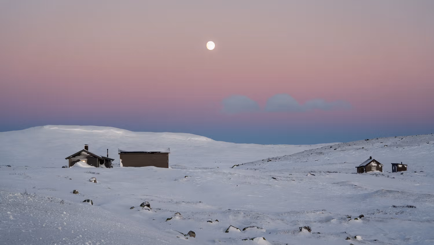 Some mountain cabins in the winter landscape underneath a pink sky with a full moon in the background.
