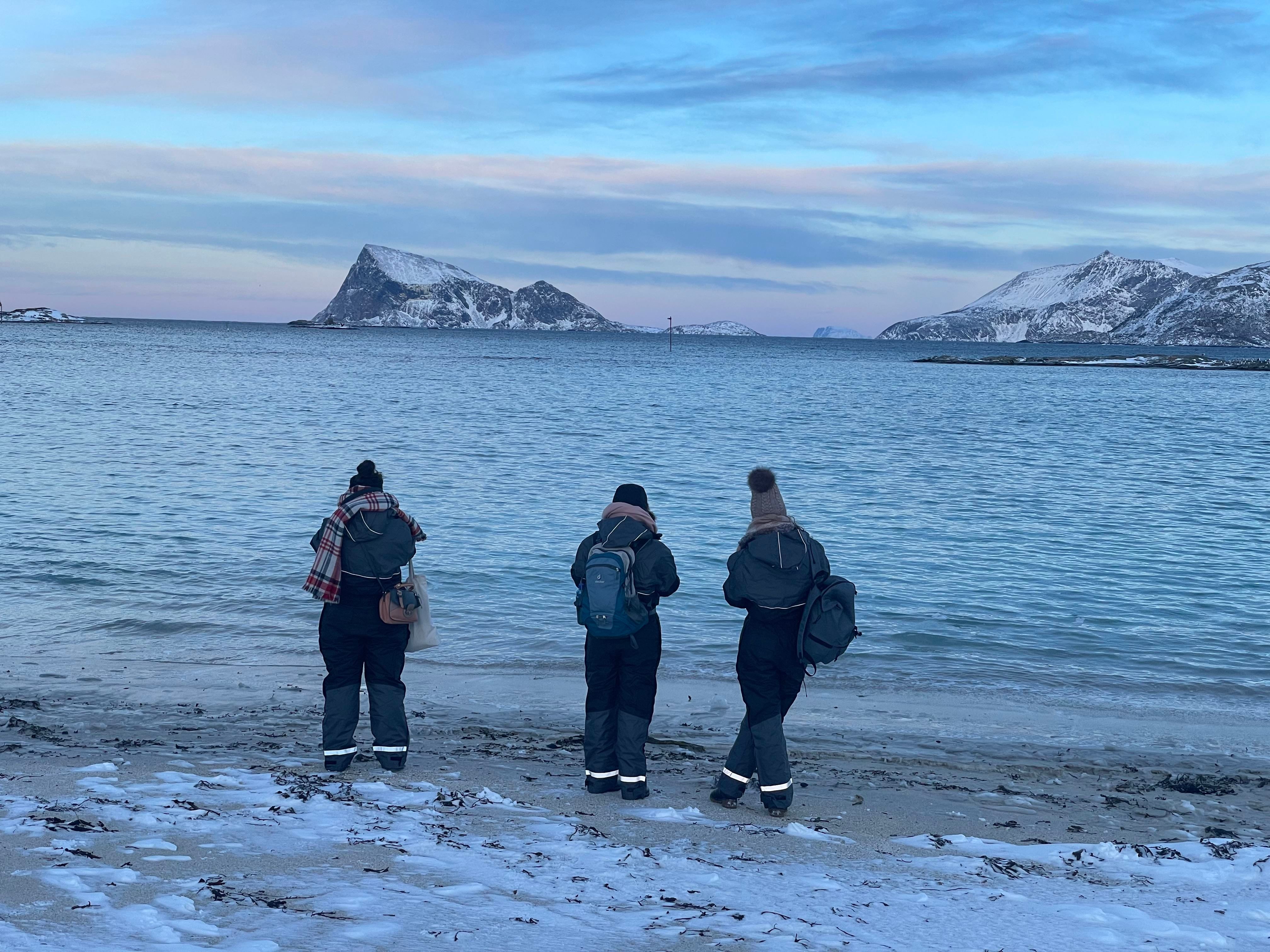 Three people on a snowy beach with fjord and mountains in the background under a blue-pink sky