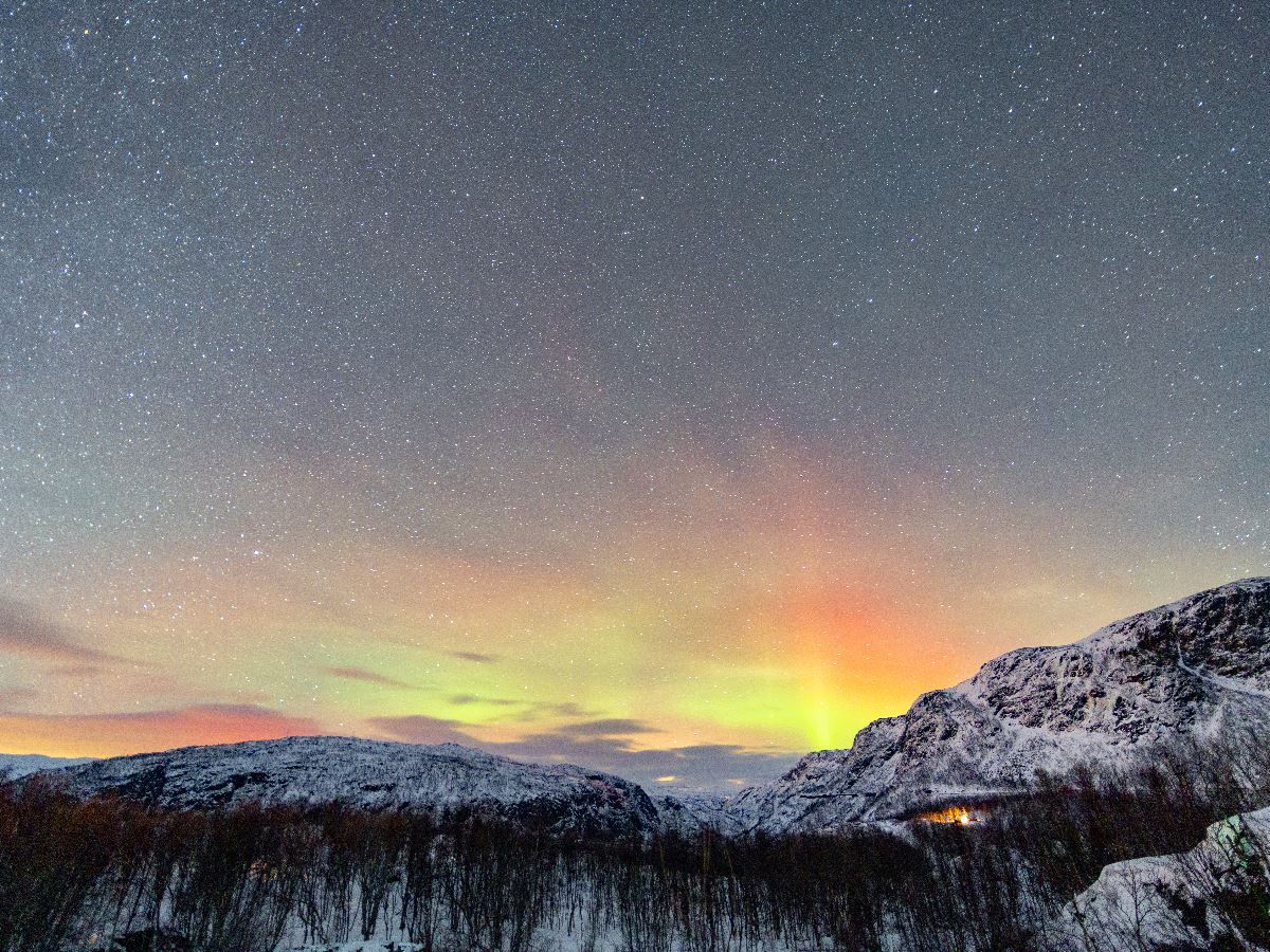Starry night sky with green and orange aurora above snow-covered mountains and a dark winter forest..