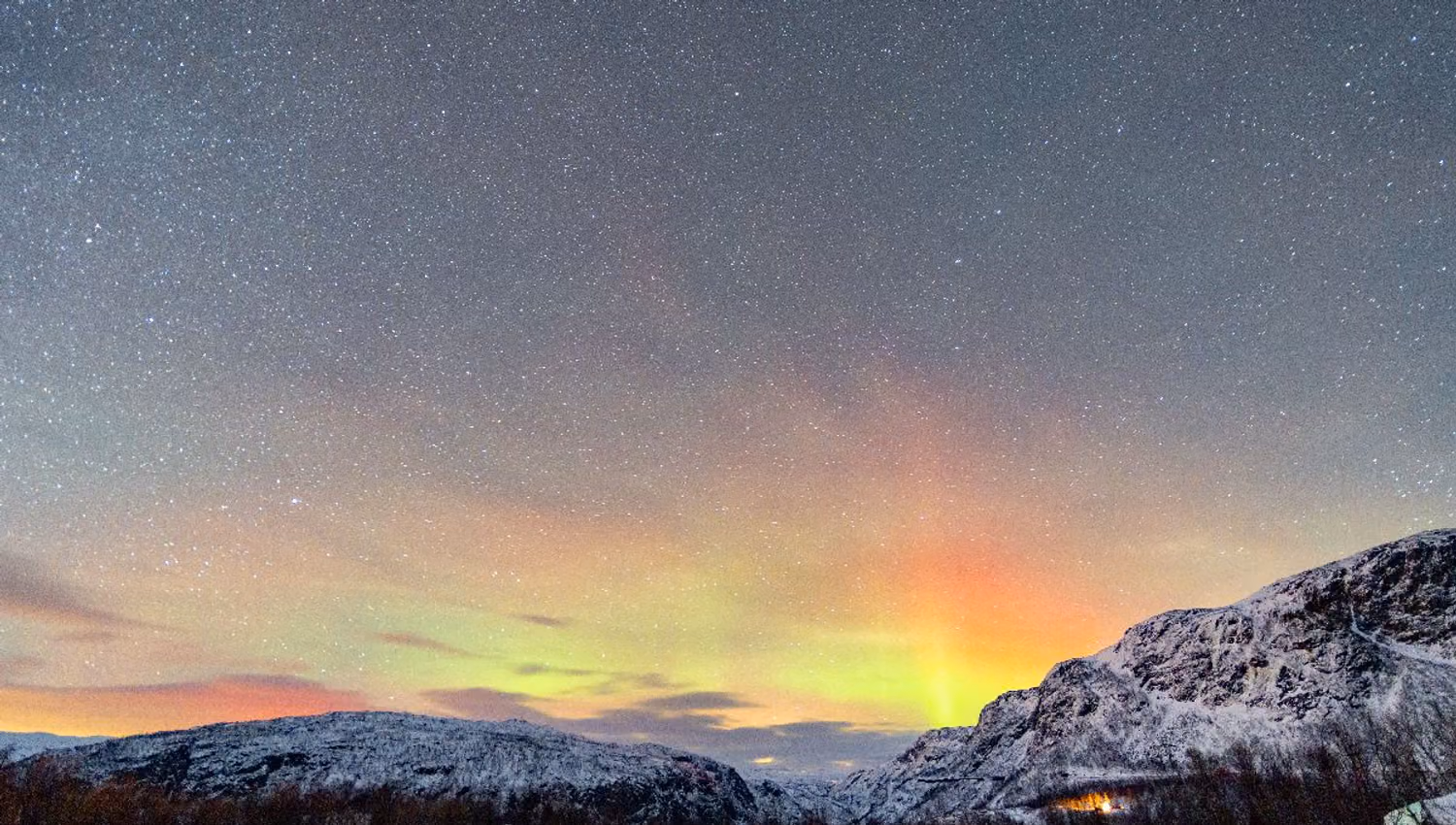 Starry night sky with green and orange aurora above snow-covered mountains and a dark winter forest..