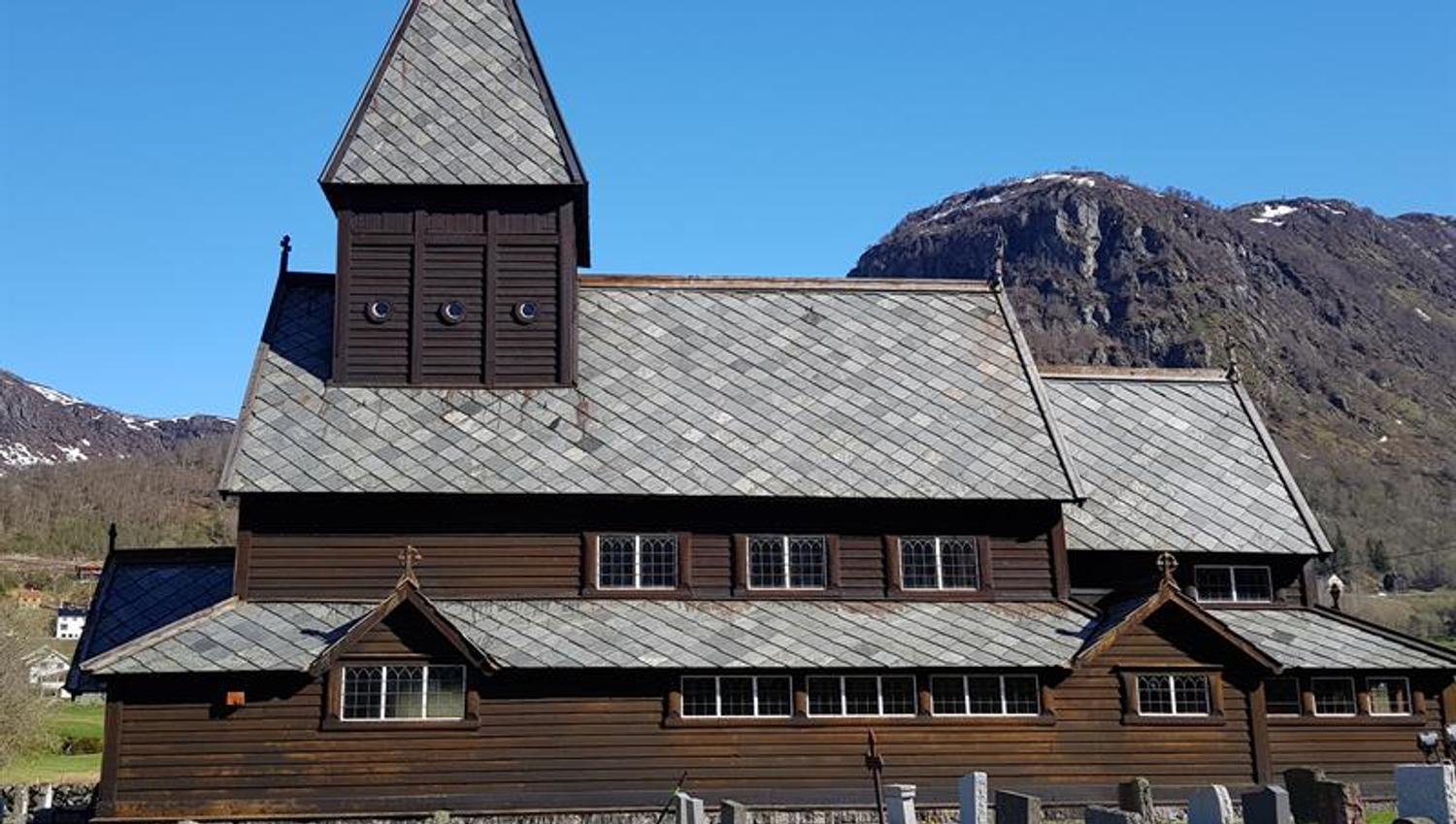 Røldal Stave Church in the spring sun with snowy mountain peaks – a beautiful meeting of culture and nature.