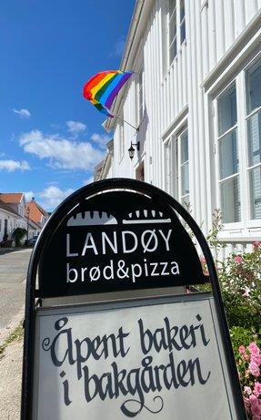 A sidewalk sign announcing that the bakery in the backyard is open. A rainbow flag is waving in the wind behind it.