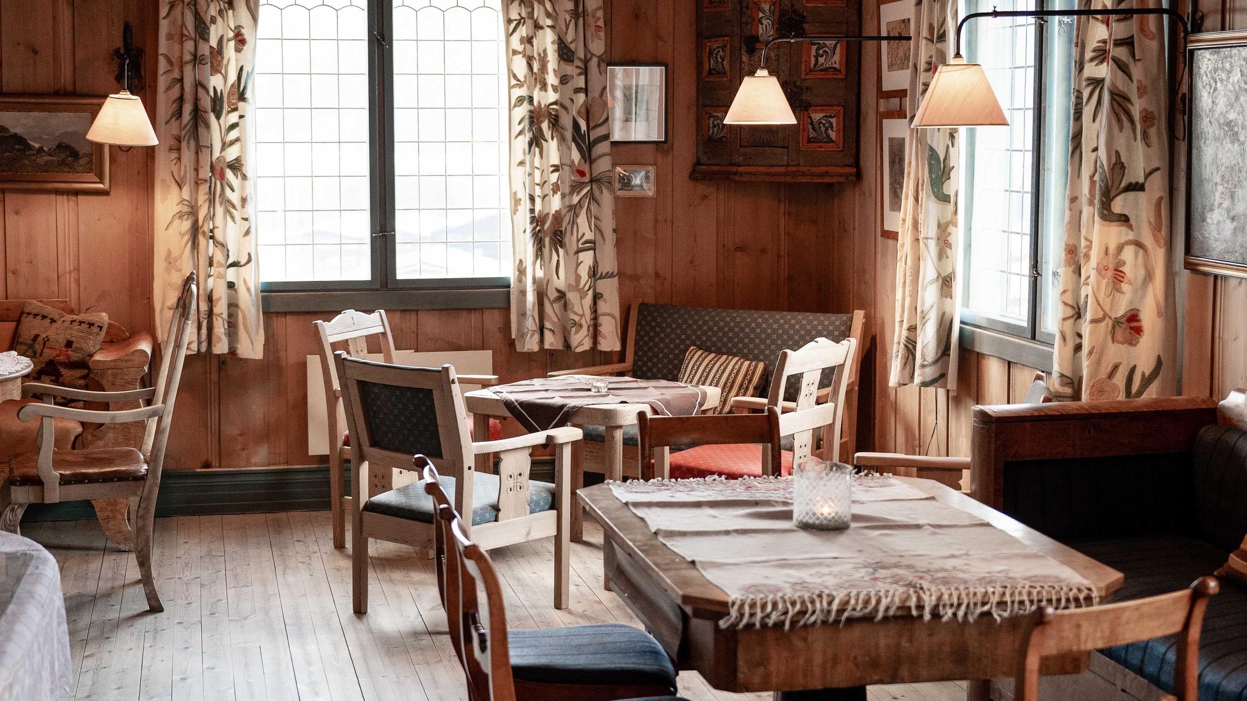 Warm wooden dining room with small tables, mixed chairs, fabric curtains, and hanging lamps by bright windows..