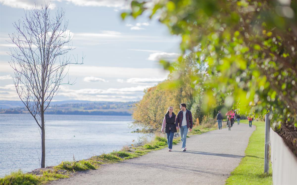 Folk går langs en gangvei ved vannet på en lys dag med trær på siden.