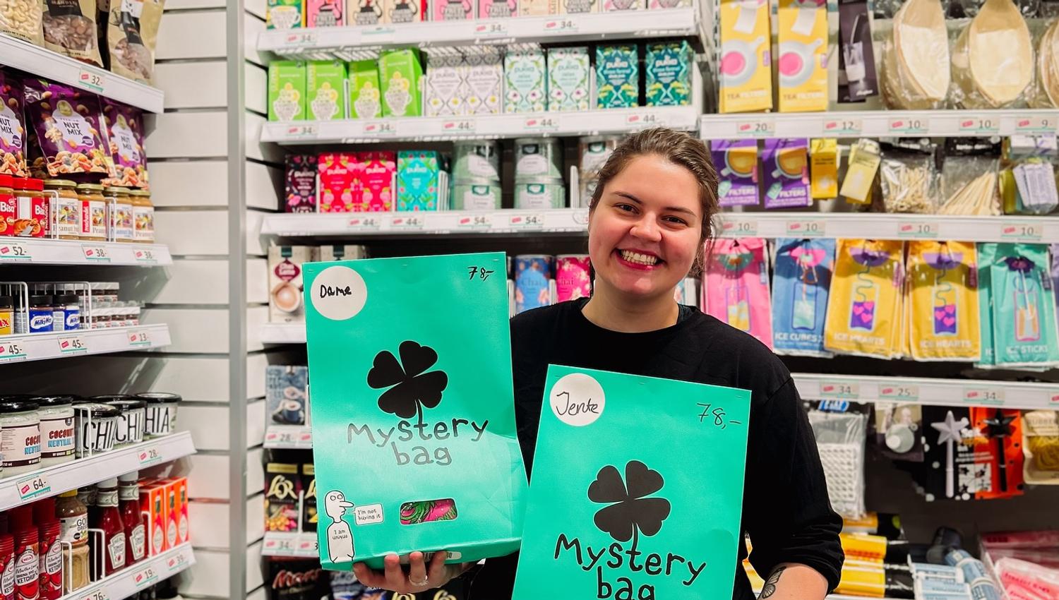 Smilende ansatt fra Normal, viser frem "Mystery Bag" foran hylle med varer