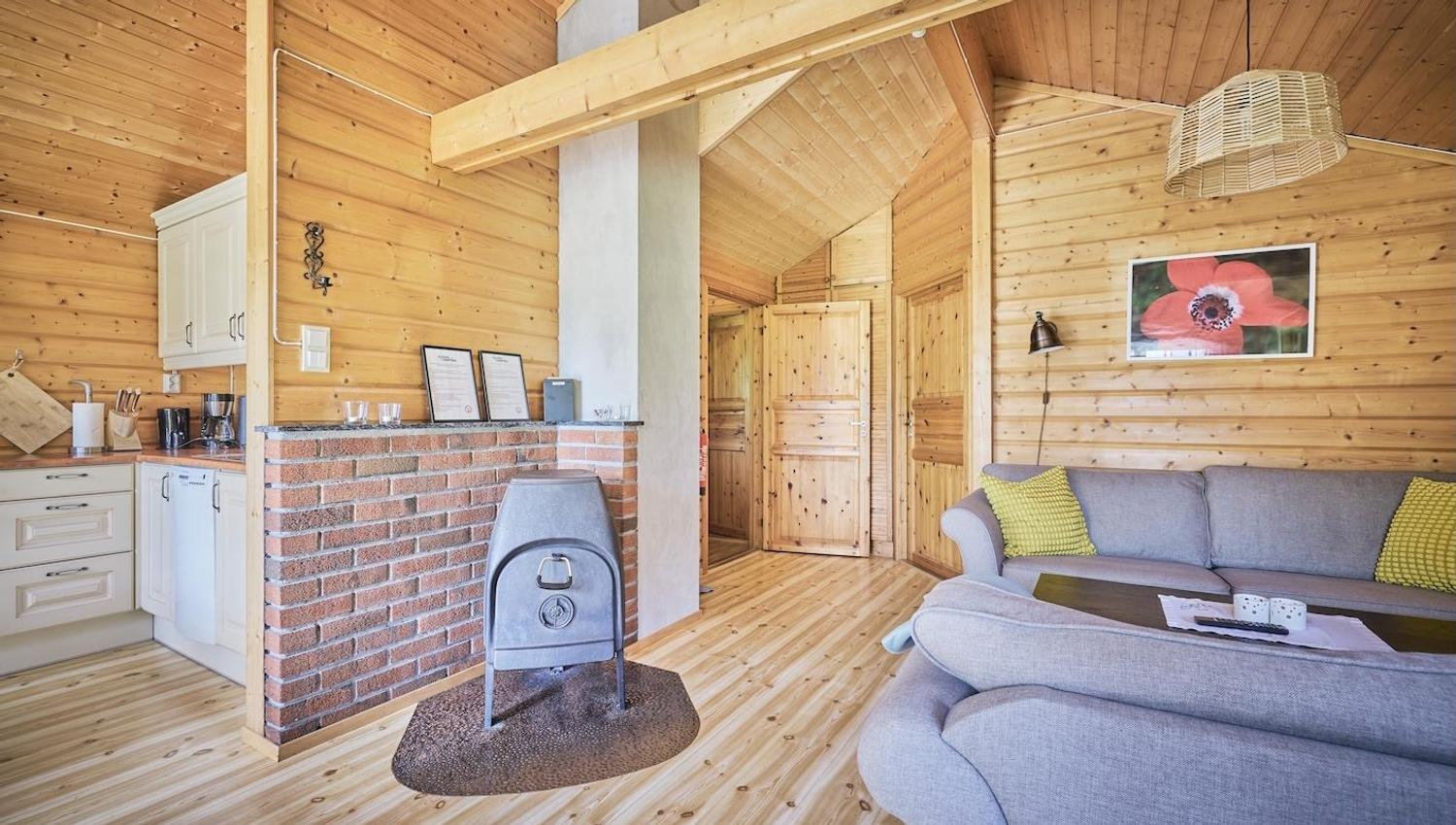 Lys hytte med trepanel, open kjøkenkrok, vedomn ved mursteinsvegg og grå sofa med puter og bilete..
