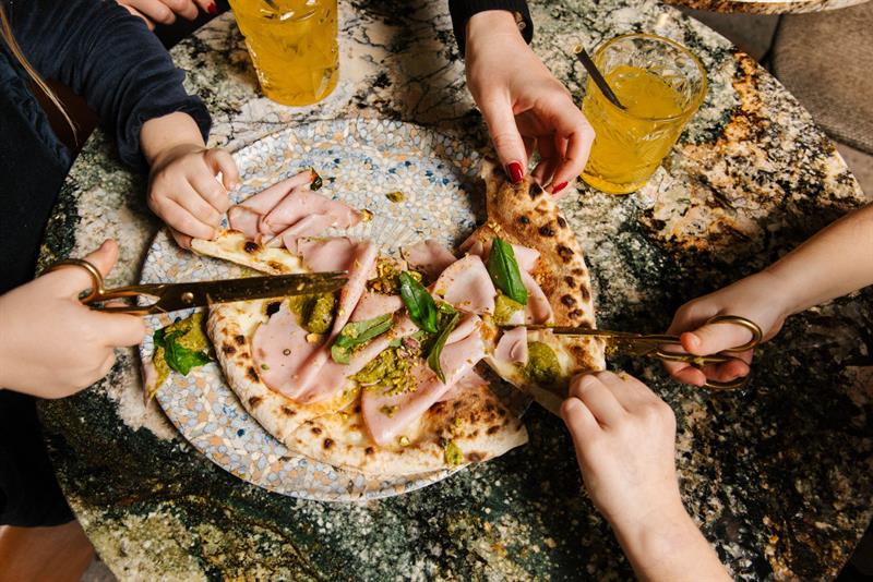 Flere hender deler en pizza toppet med mortadella og pesto ved å klippe den med saks ved et marmorbord.