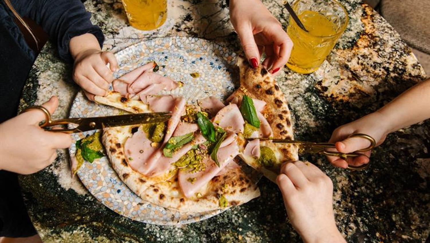Flere hender deler en pizza toppet med mortadella og pesto ved å klippe den med saks ved et marmorbord.