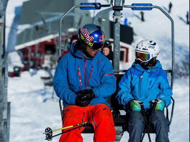 Two skiers in blue jackets sit on a chairlift holding poles, with snowy slopes and ski resort buildings behind..