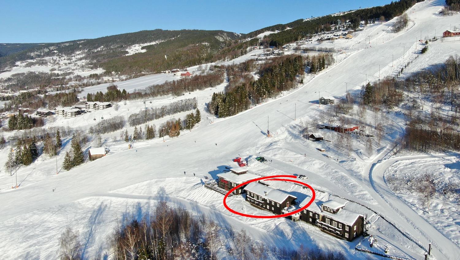 Flygvy över snötäckt skidområde med skidbacke, skog och byggnader; en stuga är markerad med röd ring.