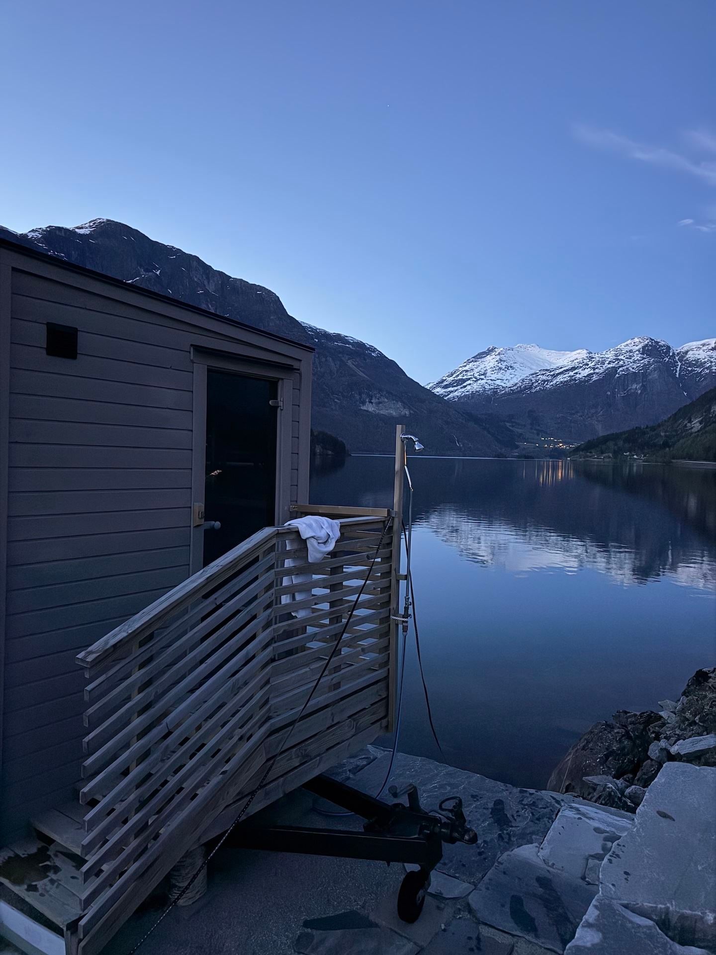 Wooden sauna deck beside a calm lake at dusk, with snow-capped mountains reflected in the water under clear sky..