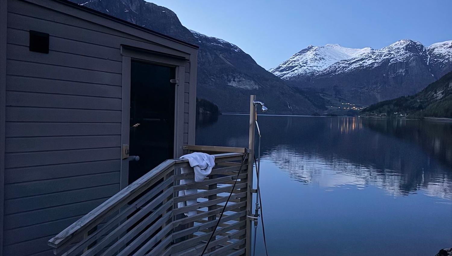 Wooden sauna deck beside a calm lake at dusk, with snow-capped mountains reflected in the water under clear sky..