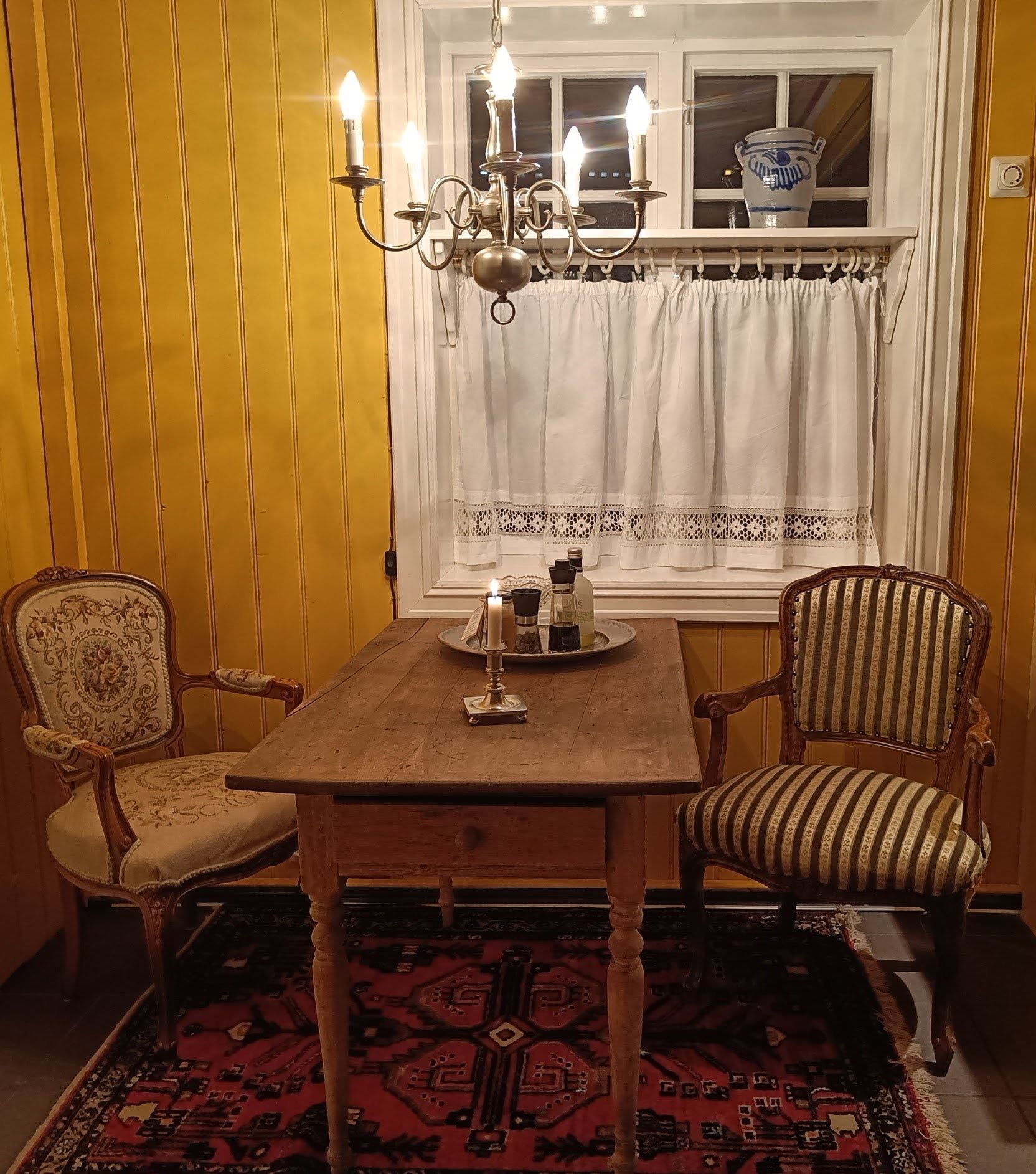 Orheimstunet in Nordfjord - Small dining area with wooden table, two vintage armchairs, chandelier and candle, beneath a curtained window..