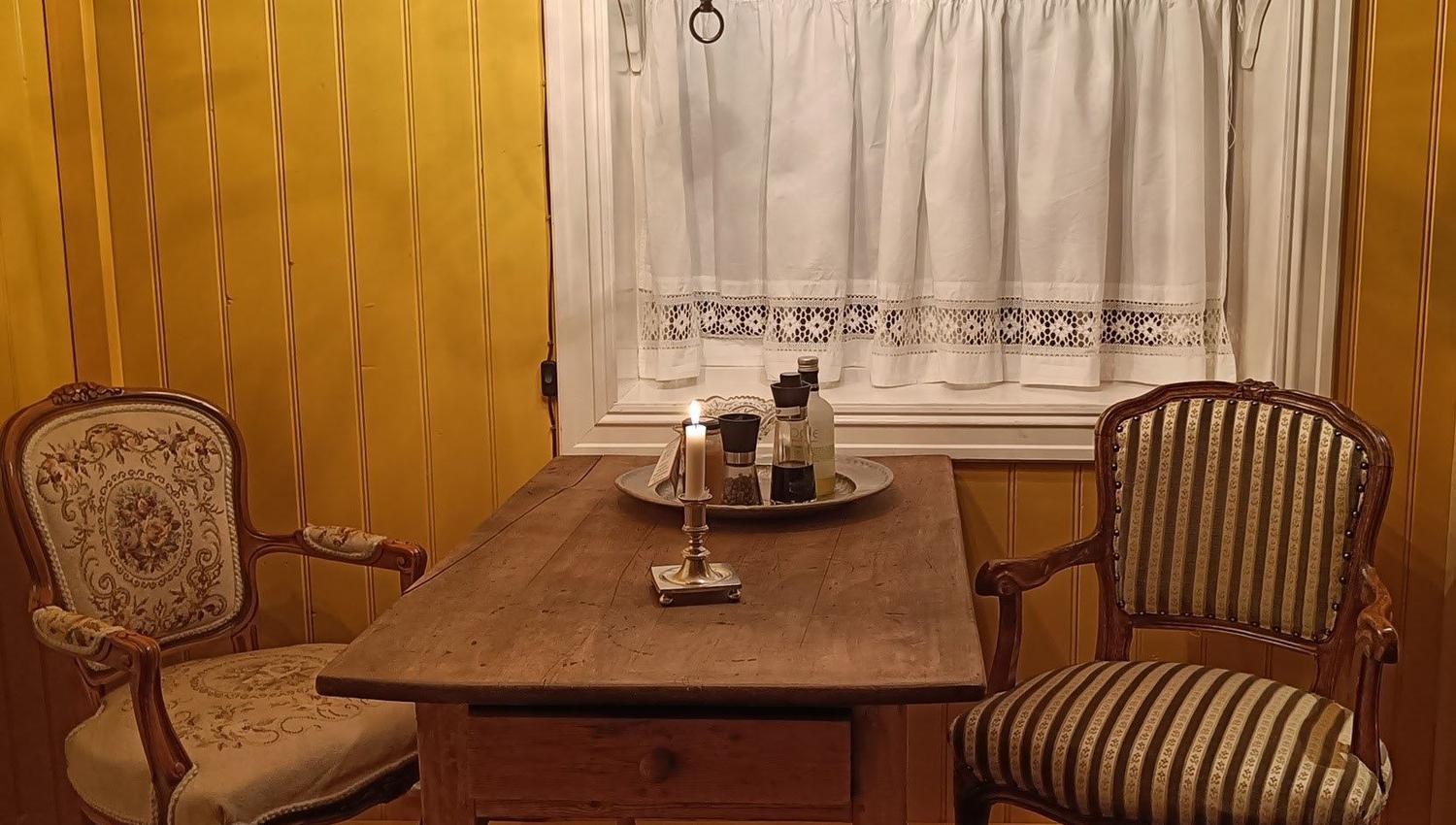 Orheimstunet in Nordfjord - Small dining area with wooden table, two vintage armchairs, chandelier and candle, beneath a curtained window..