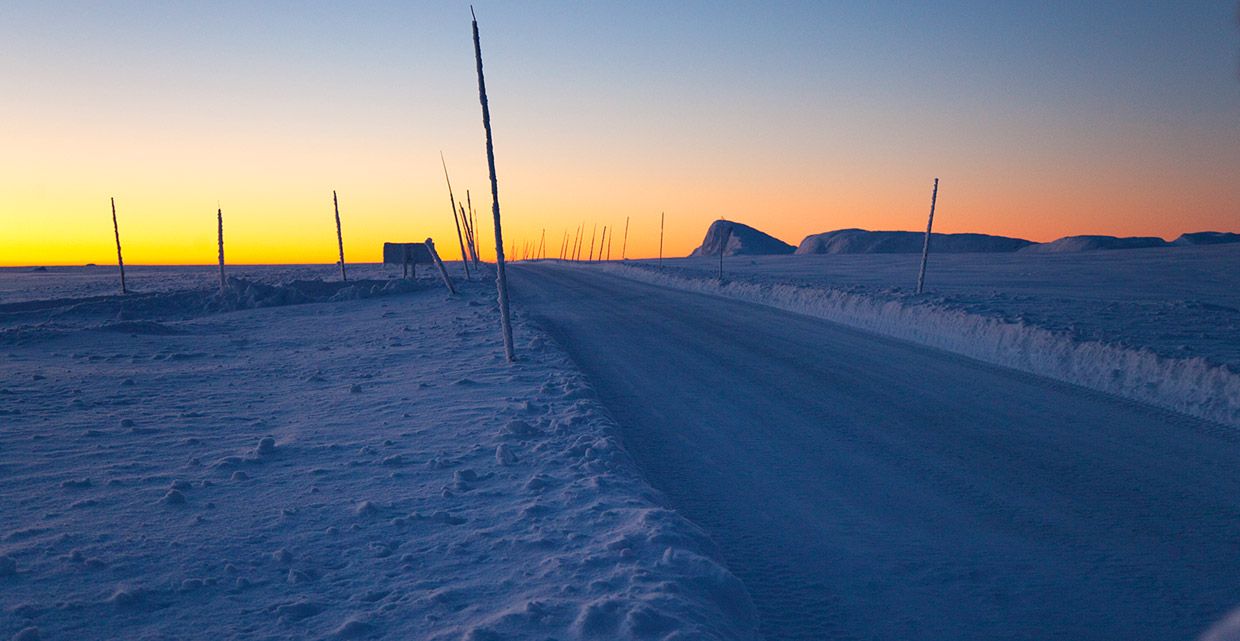 A snowy road after sunset in the beginning twilight as the remaining light from the sun colour the horizon yellow-orange.