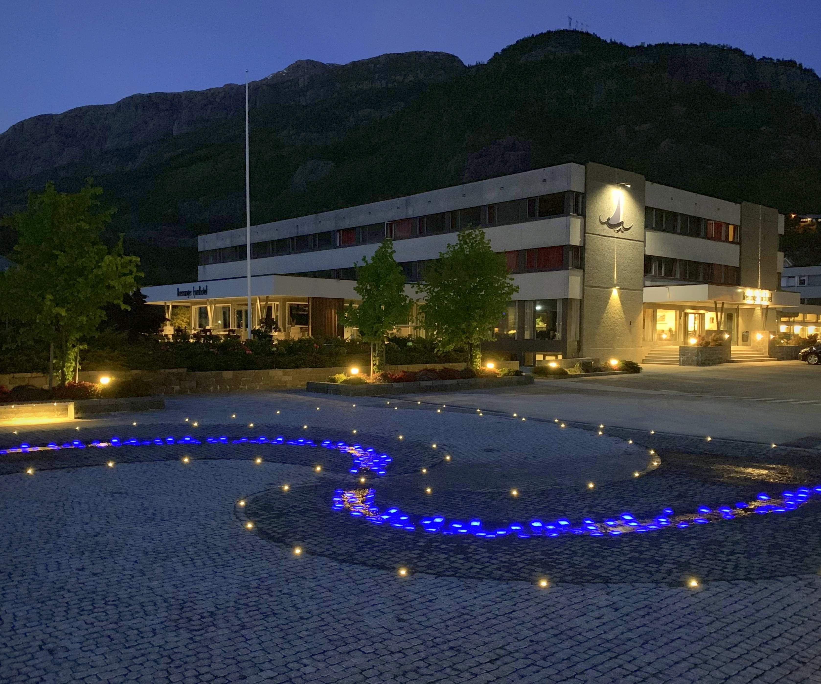 Opplyst hotellbygg ved foten av fjell i skumring, med steinlagt plass og blå lys i mønster i forgrunnen..