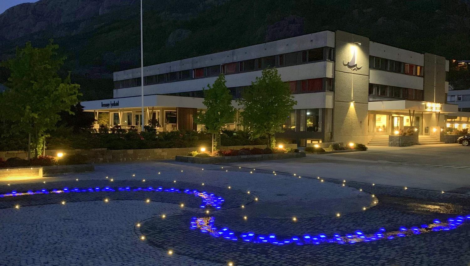Opplyst hotellbygg ved foten av fjell i skumring, med steinlagt plass og blå lys i mønster i forgrunnen..