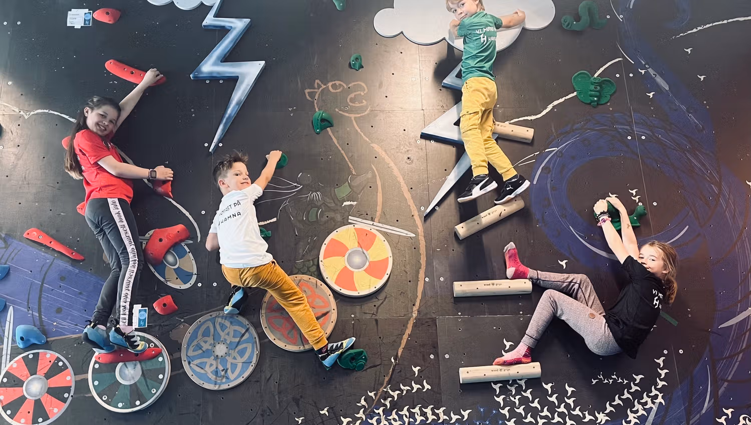 Four children climb a colorful indoor bouldering wall with holds, cloud and lightning shapes, and shield art..