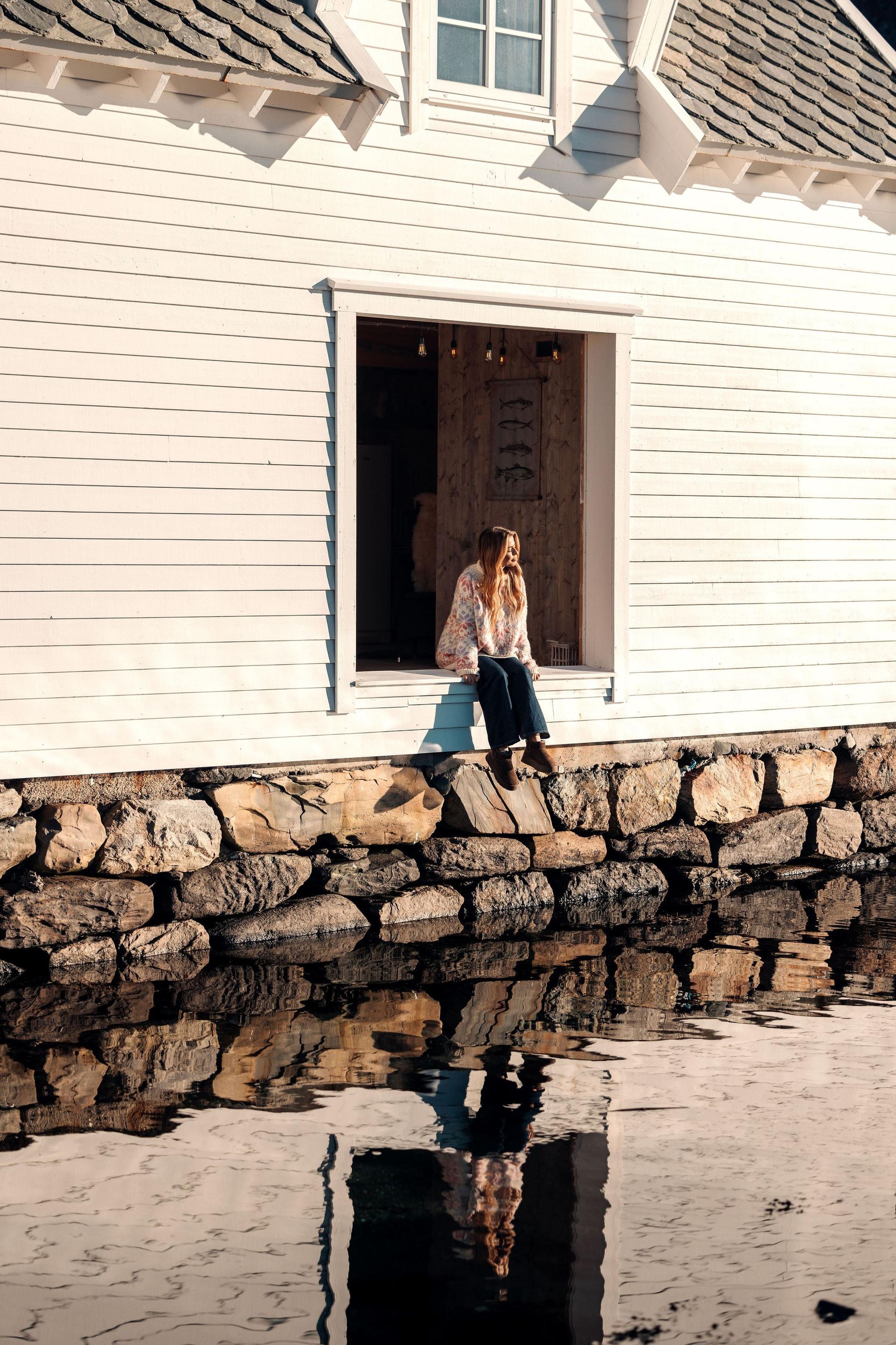 A person sits in a large open window of a white wooden building above a stone wall, reflected in calm water at the Seahouse at Hornelen Panorama in Bremanger..