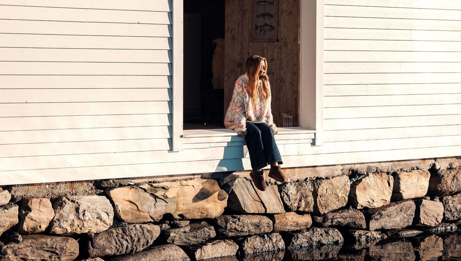 A person sits in a large open window of a white wooden building above a stone wall, reflected in calm water at the Seahouse at Hornelen Panorama in Bremanger..