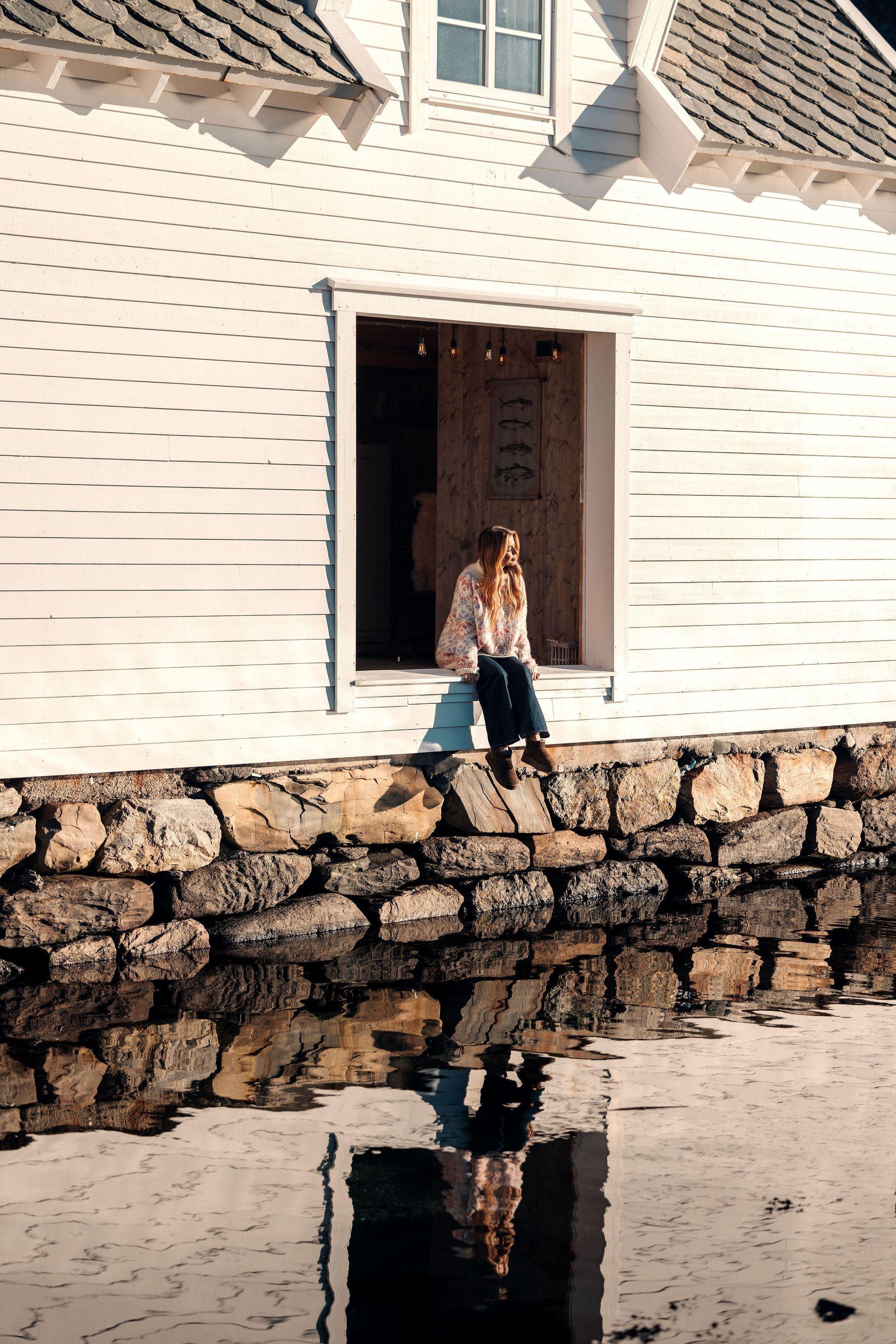 A person sits in a large open window of a white wooden building above a stone wall, reflected in calm water at the Seahouse at Hornelen Panorama in Bremanger..