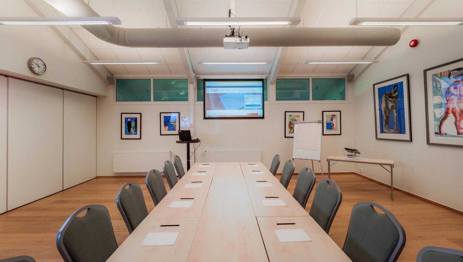 Bright conference room with long tables, chairs, notepads, projector screen, flip chart, and framed art.