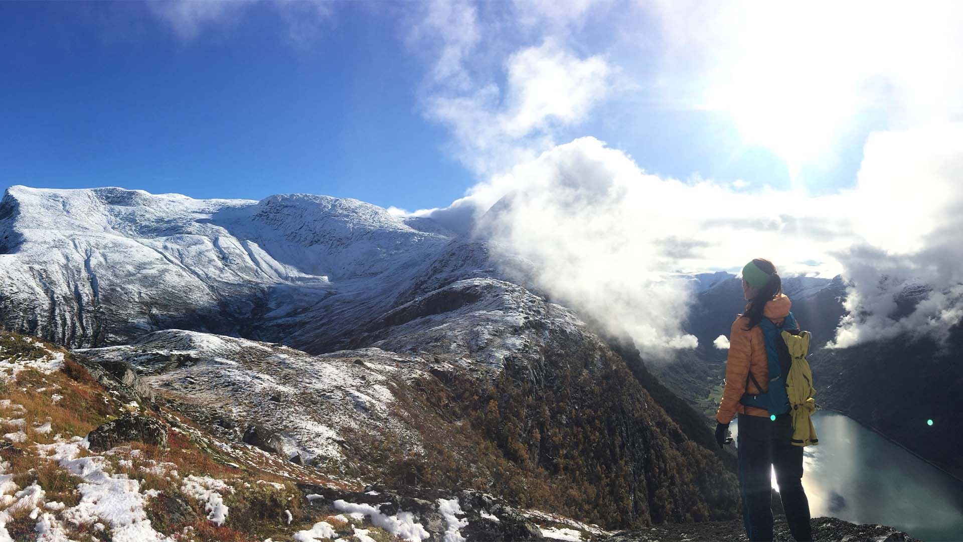 Hiker with backpack stands on a snowy ridge, looking over a mountain valley with clouds and a lake below..