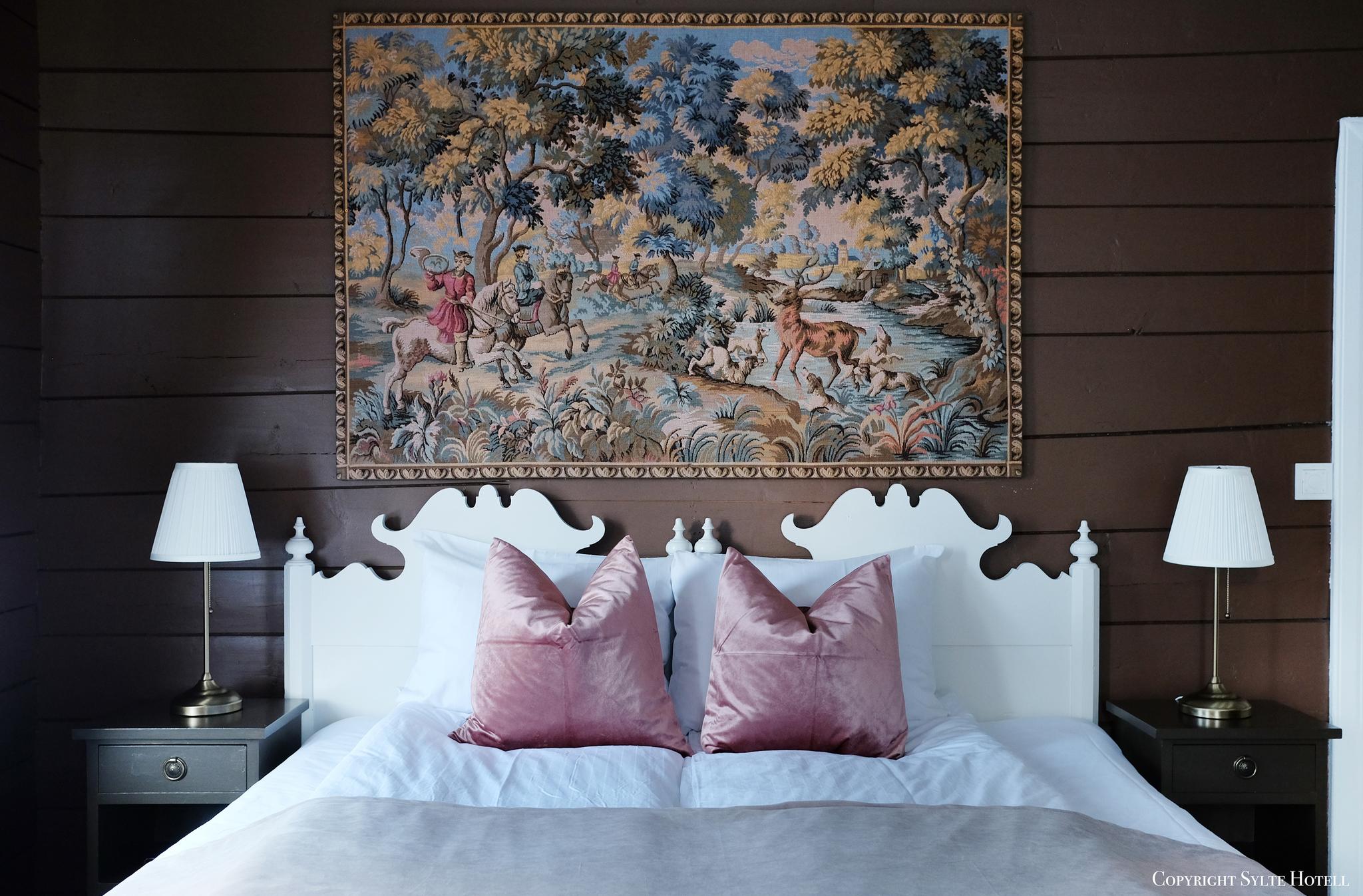 A bed in shades of pink, with a large embroidery above the bed with a historical motif.