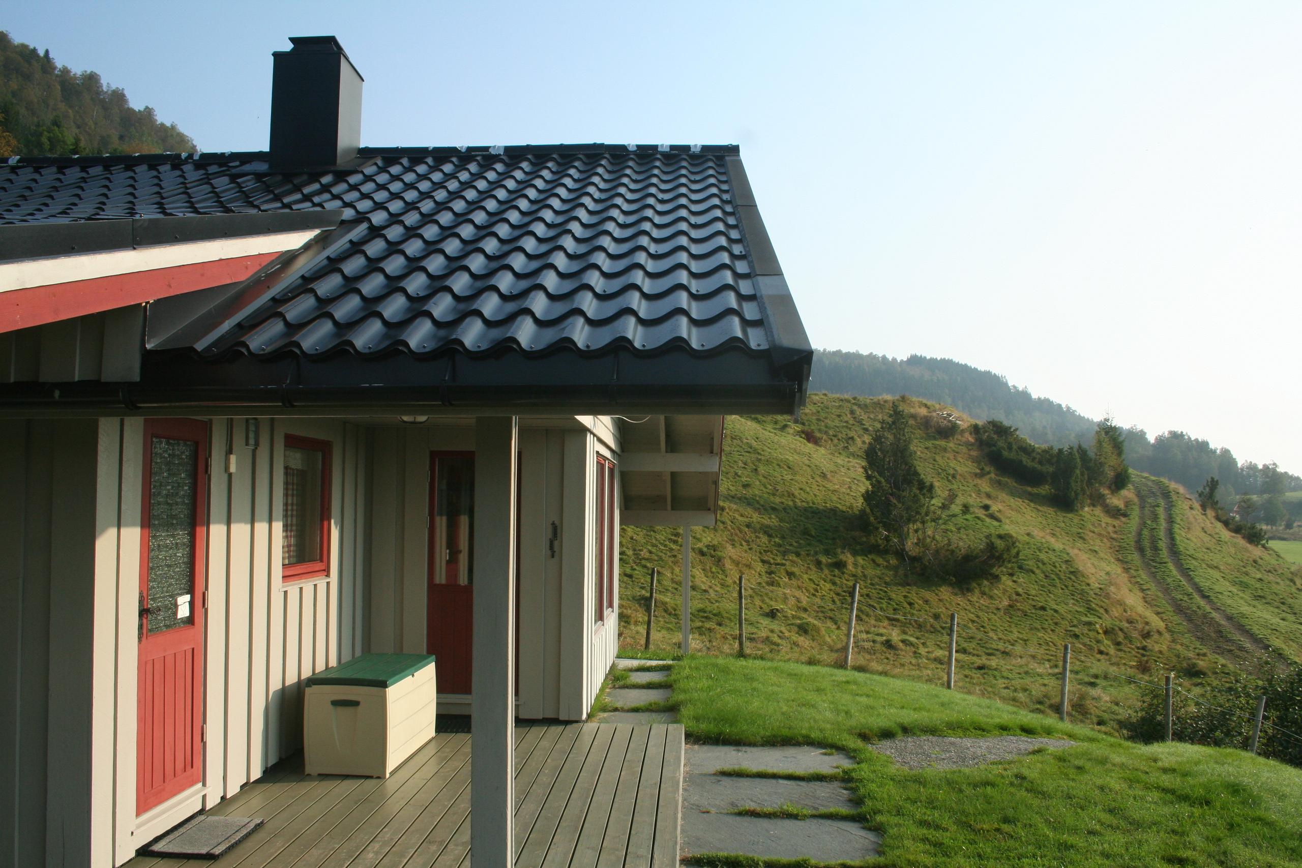 Traditional Norwegian cabin with a covered terrace, red doors, and a scenic view of the green hills in Hardanger.