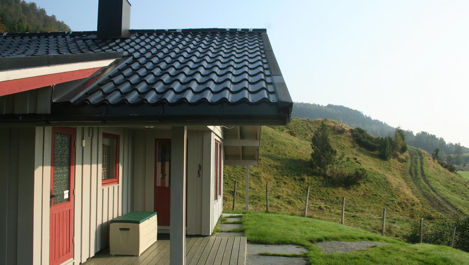 Traditional Norwegian cabin with a covered terrace, red doors, and a scenic view of the green hills in Hardanger.