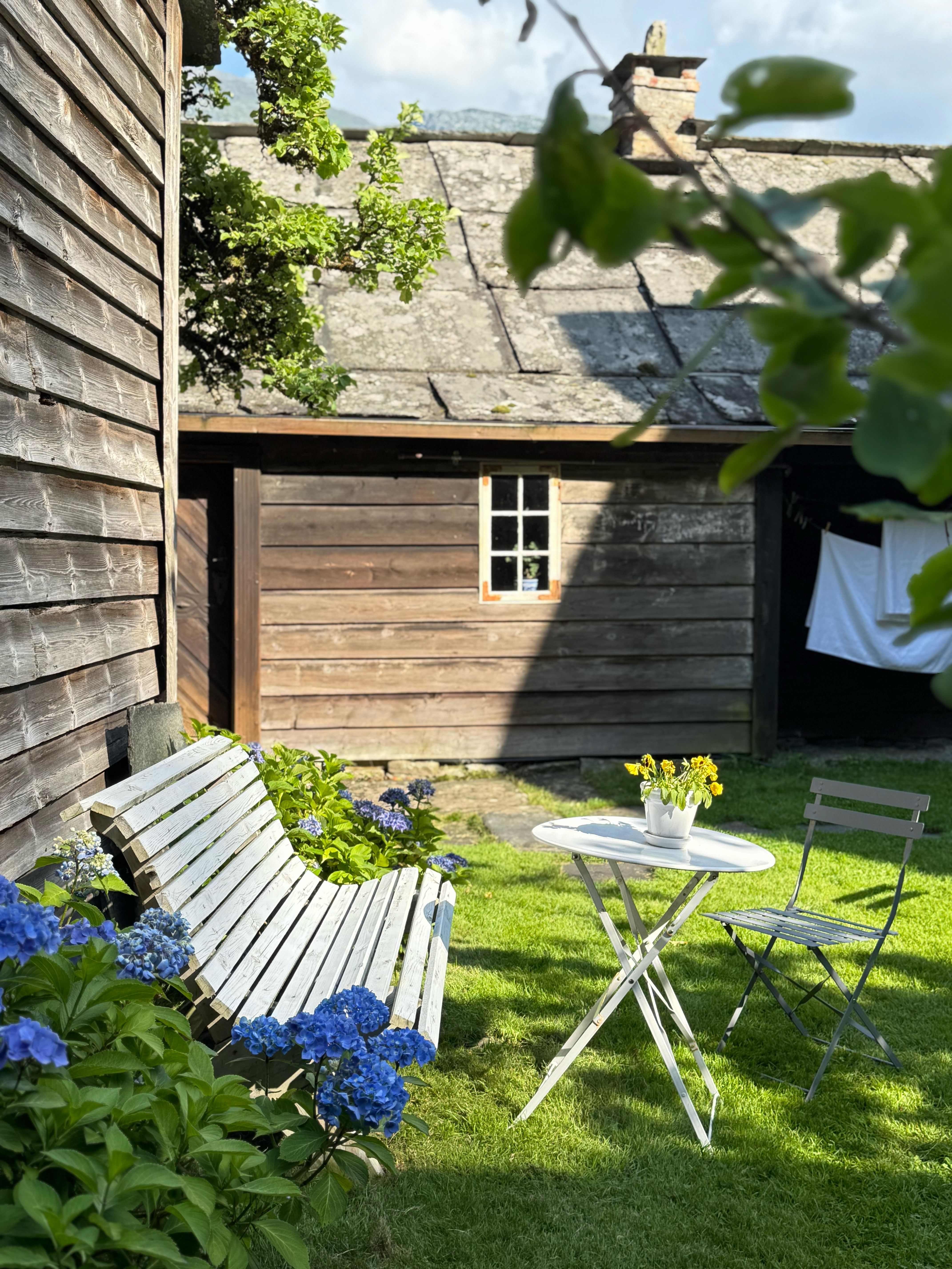 A small garden corner in Agatunet with a white-painted bench, a small table with flowers, and a chair, surrounded by blue hydrangeas in front of an ol