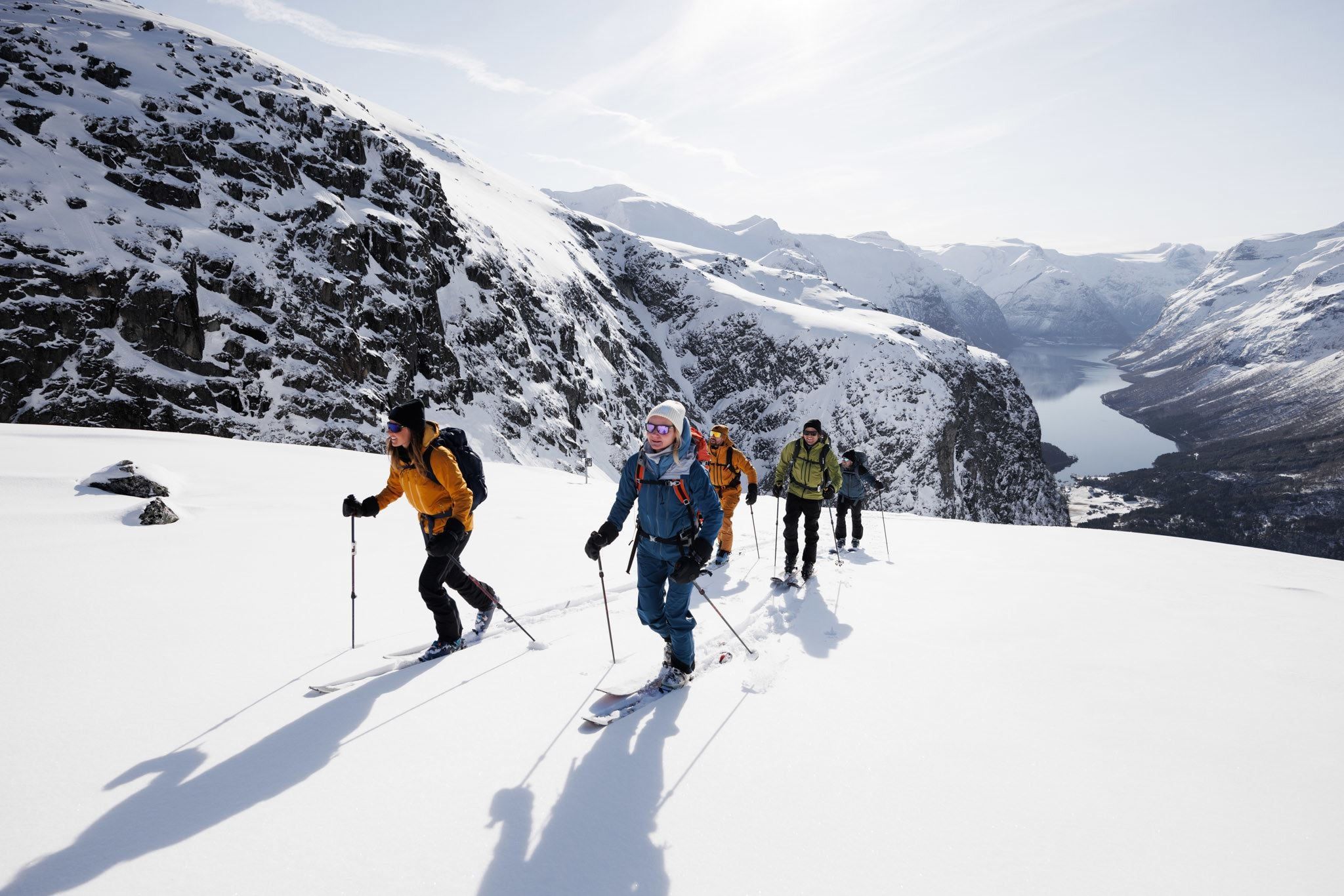 A group of skiers hike uphill on a snowy ridge with poles, with steep rocky mountains and a fjord below.