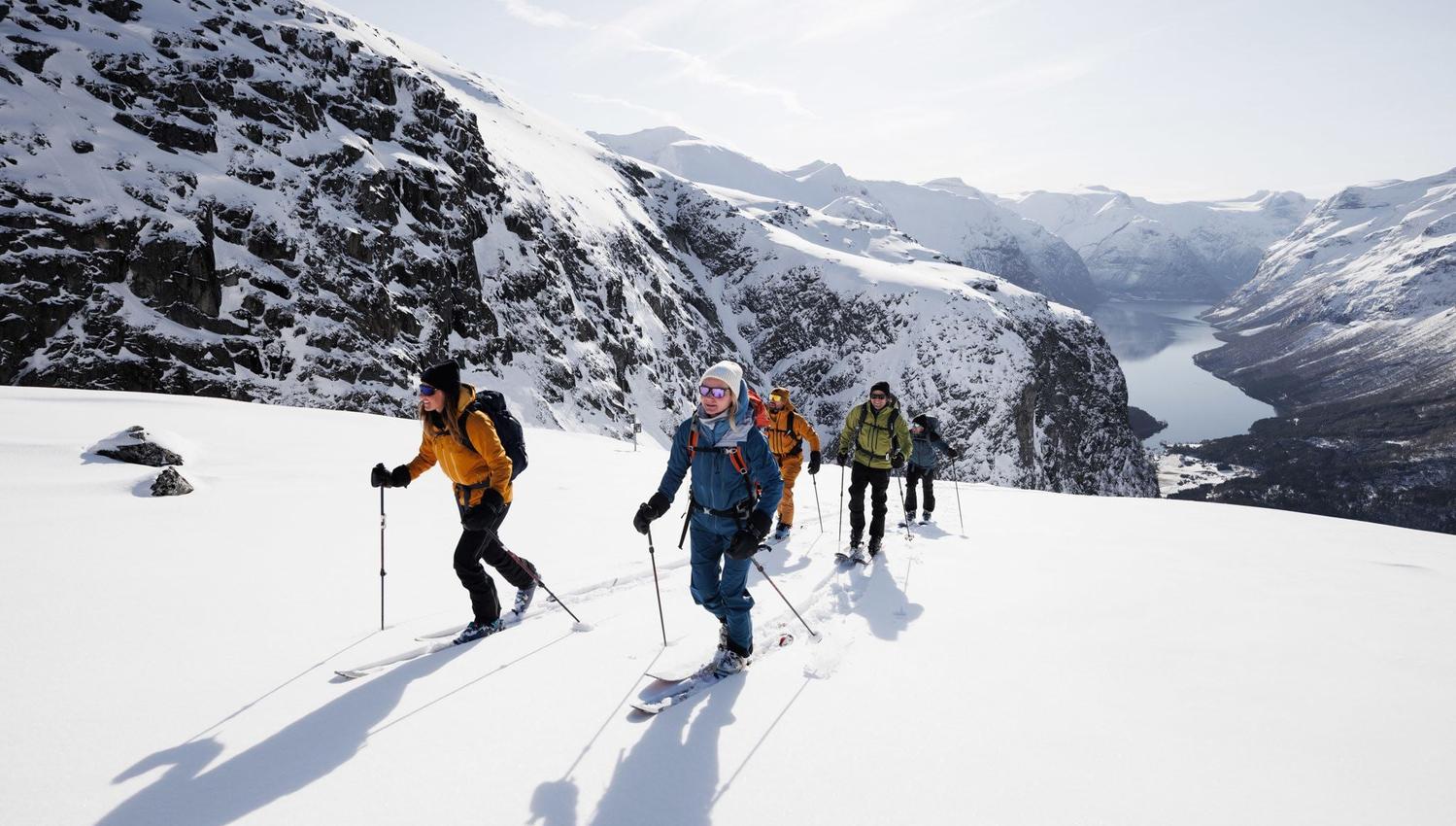 A group of skiers hike uphill on a snowy ridge with poles, with steep rocky mountains and a fjord below.