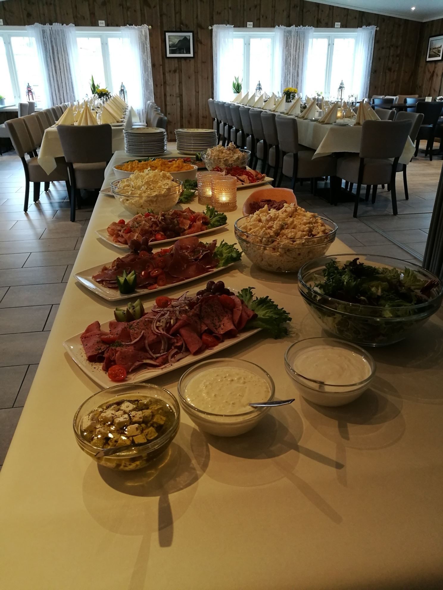 Long buffet table with salads, meats, dips, and stacked plates in a rustic dining room with set tables and chairs.