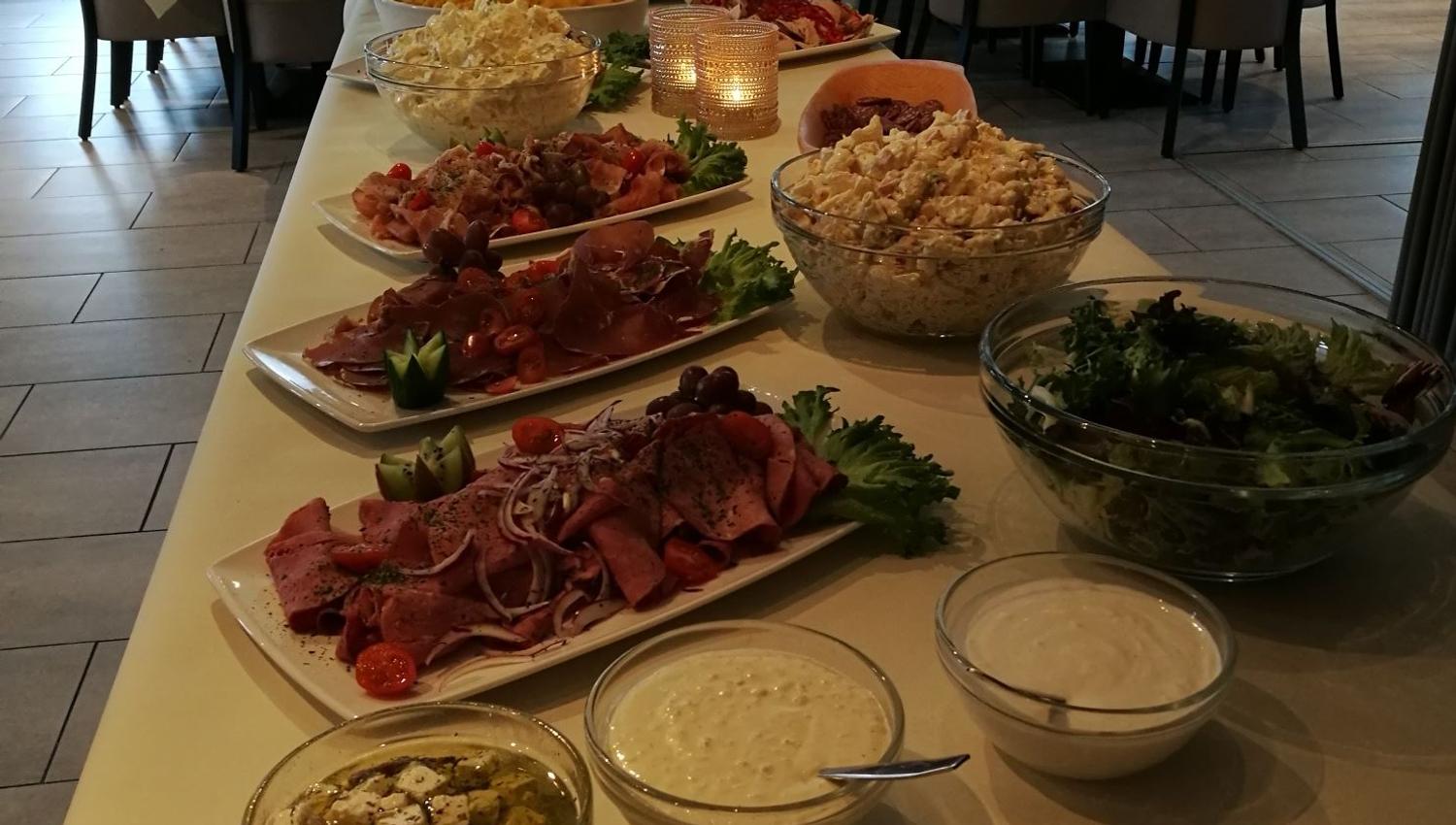 Long buffet table with salads, meats, dips, and stacked plates in a rustic dining room with set tables and chairs.