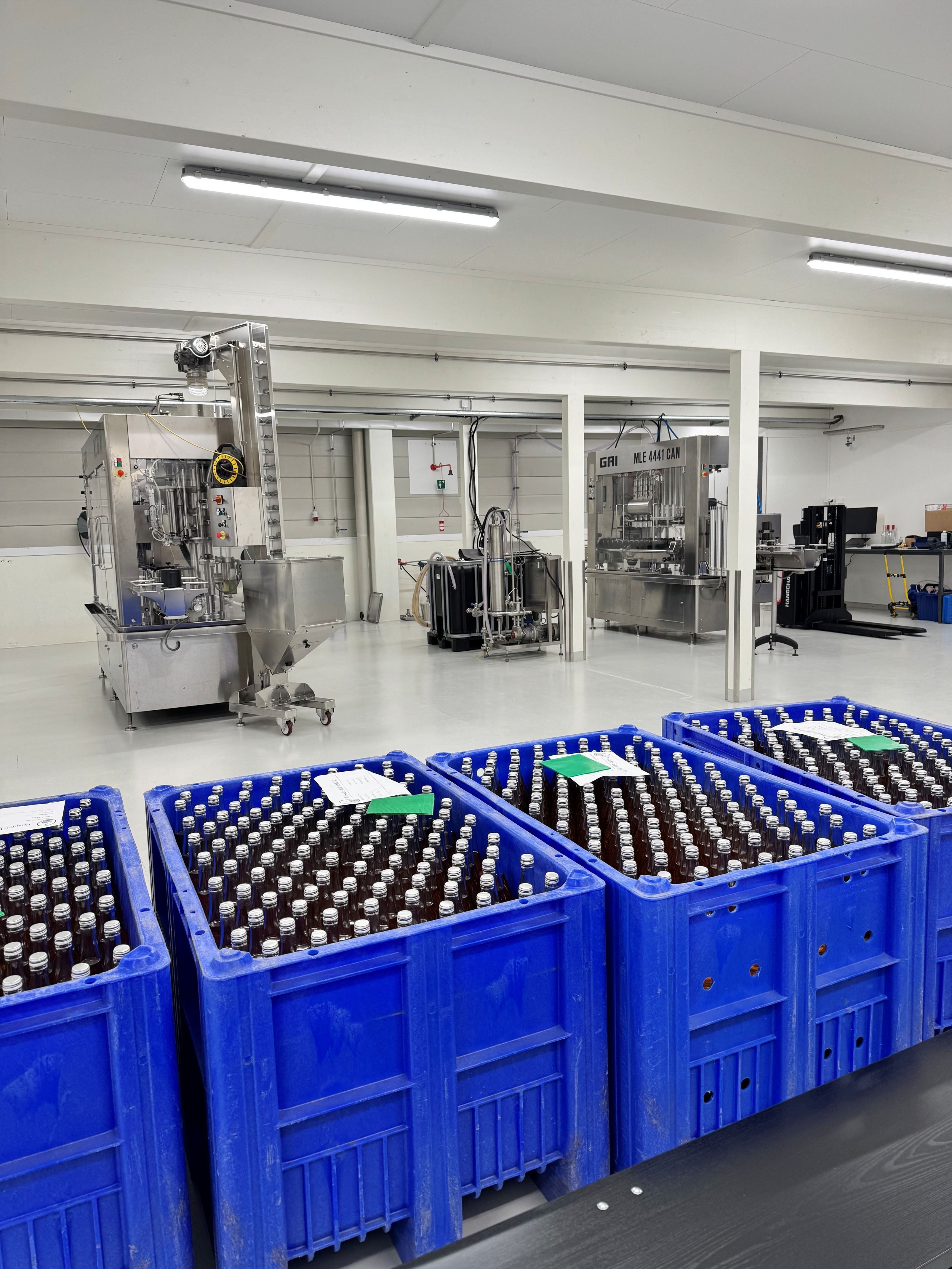 Blue plastic crates filled with capped glass bottles in a clean industrial room with stainless machinery and bright lights.