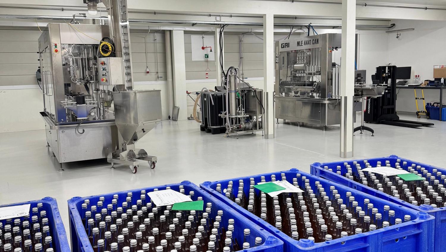 Blue plastic crates filled with capped glass bottles in a clean industrial room with stainless machinery and bright lights.