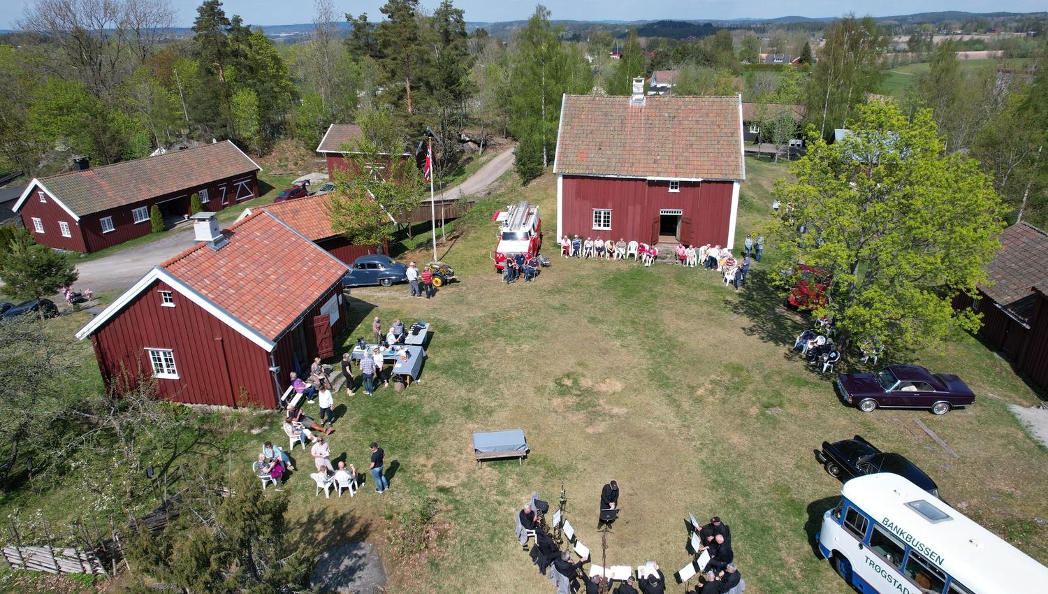Et bilde av røde gårdshus, med mennesker og gamle biler på plenen rundt.